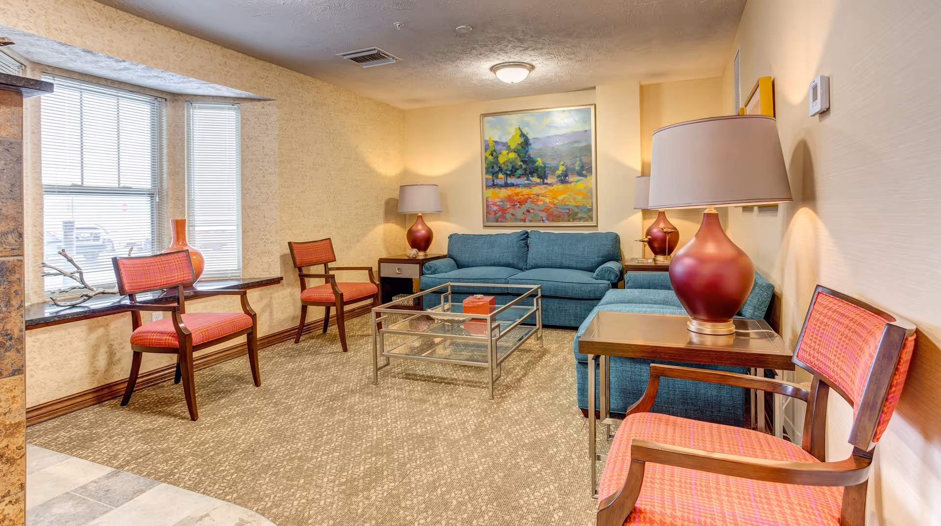 A cozy living room area with a teal sofa, two matching armchairs, and two wooden chairs with red cushions. There are two side tables with red lamps, a glass coffee table, a large window with blinds, and a colorful landscape painting on the wall.