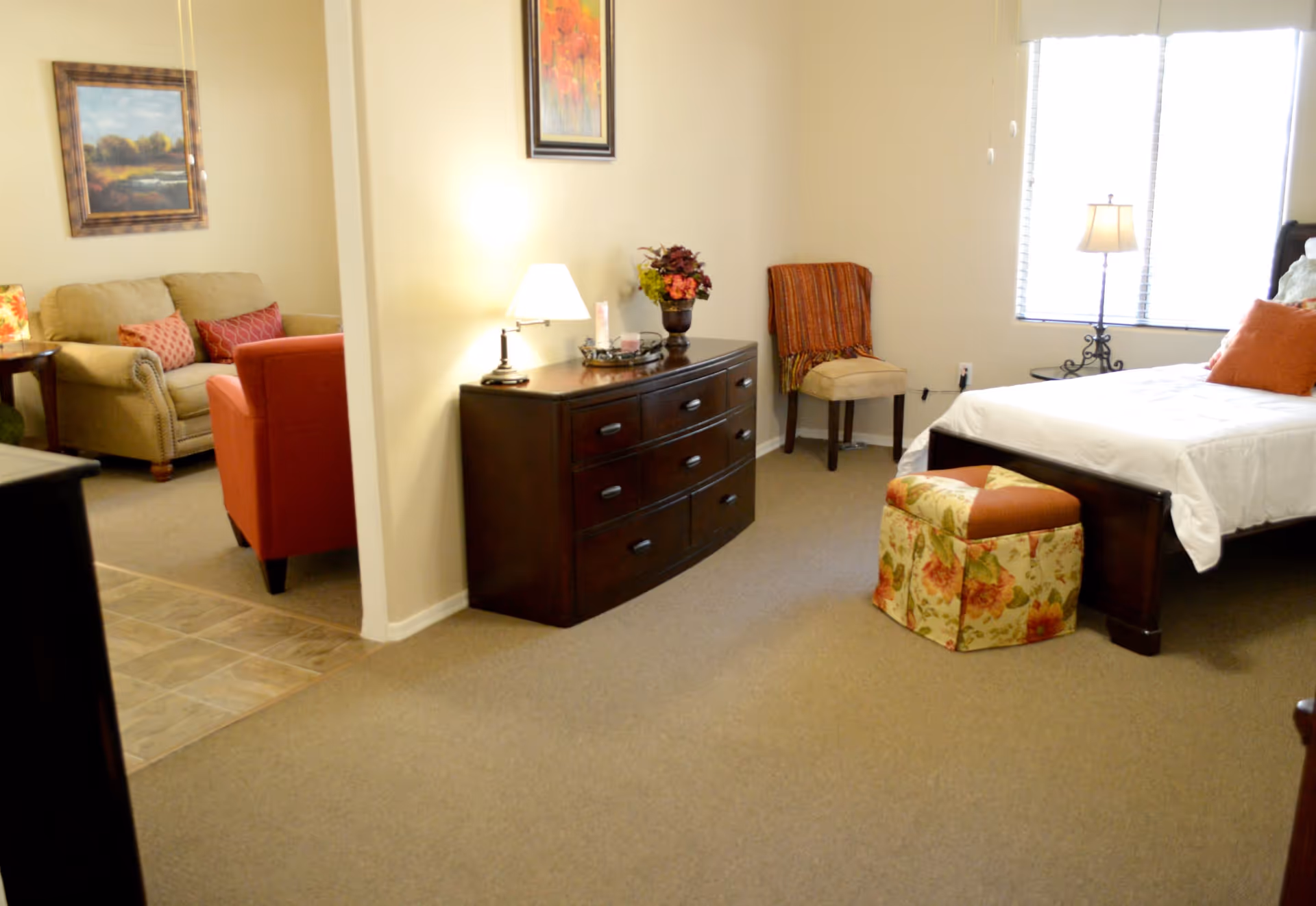 Bright assisted living suite showing a bed, dresser, chair, and a small seating area with a sofa and armchair.