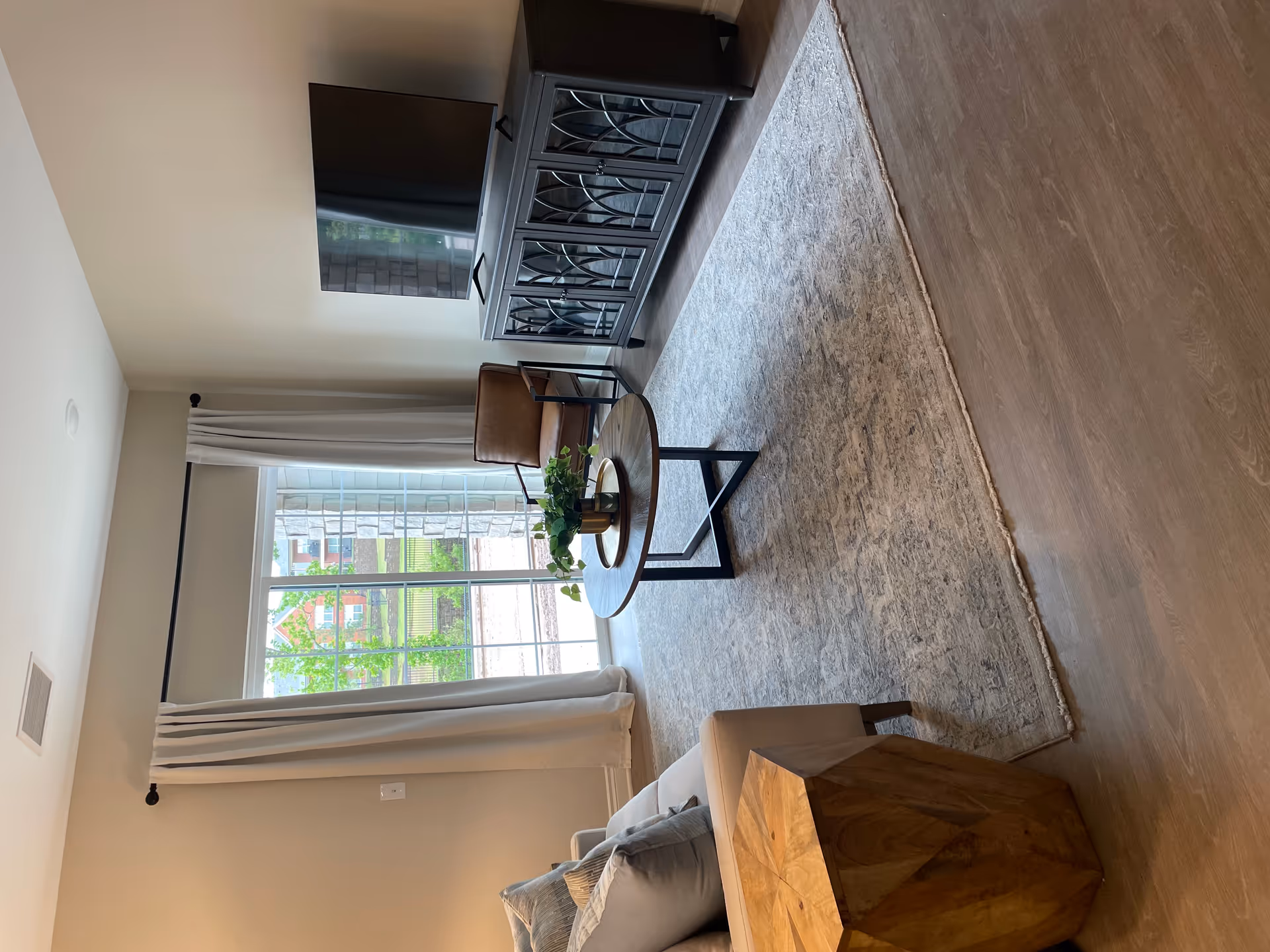 A cozy living room with a beige sofa adorned with pillows on the left, a geometric wooden side table, a round wooden coffee table with a plant centerpiece, a brown leather armchair, and a dark wooden TV stand with a flat-screen TV. The room has light-colored walls, a large window with white curtains, and wood flooring with a large area rug.