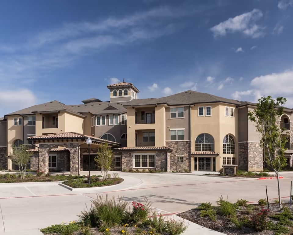 Front exterior of a three-story senior living building with stone accents, arched windows, a covered entrance, and a landscaped driveway.