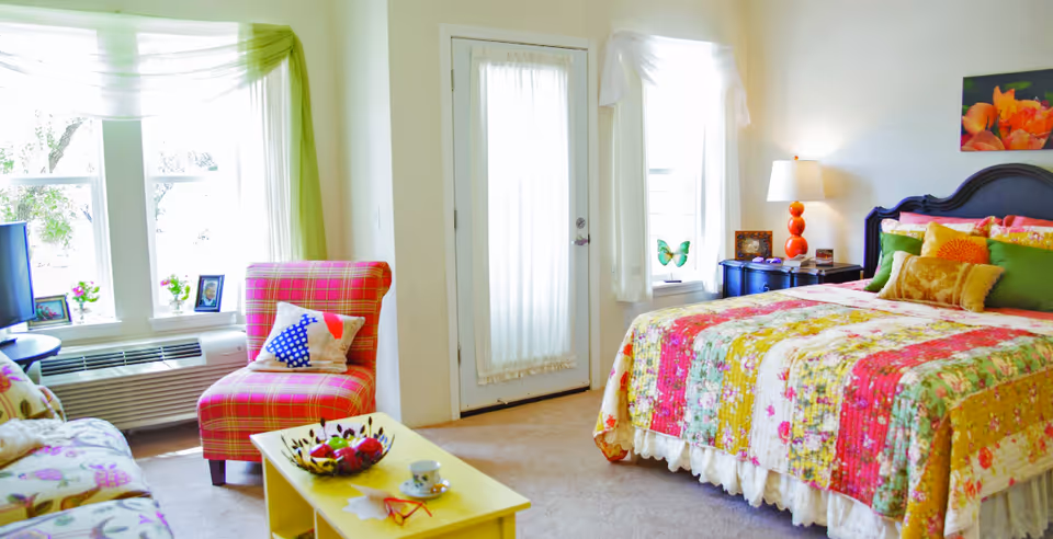 Bright and cozy bedroom with a colorful floral quilt on the bed, green and yellow pillows, a bedside table with a lamp, a pink plaid armchair with a decorative pillow, a yellow coffee table with a bowl of fruit and a cup, and large windows with sheer white and green curtains letting in natural light.