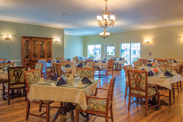 A spacious dining room with multiple tables covered in beige tablecloths, each set with glassware, napkins folded in a triangular shape, and floral centerpieces. The chairs have floral upholstery and wooden frames. The room features wooden flooring, soft lighting from chandeliers and wall sconces, and large windows letting in natural light.