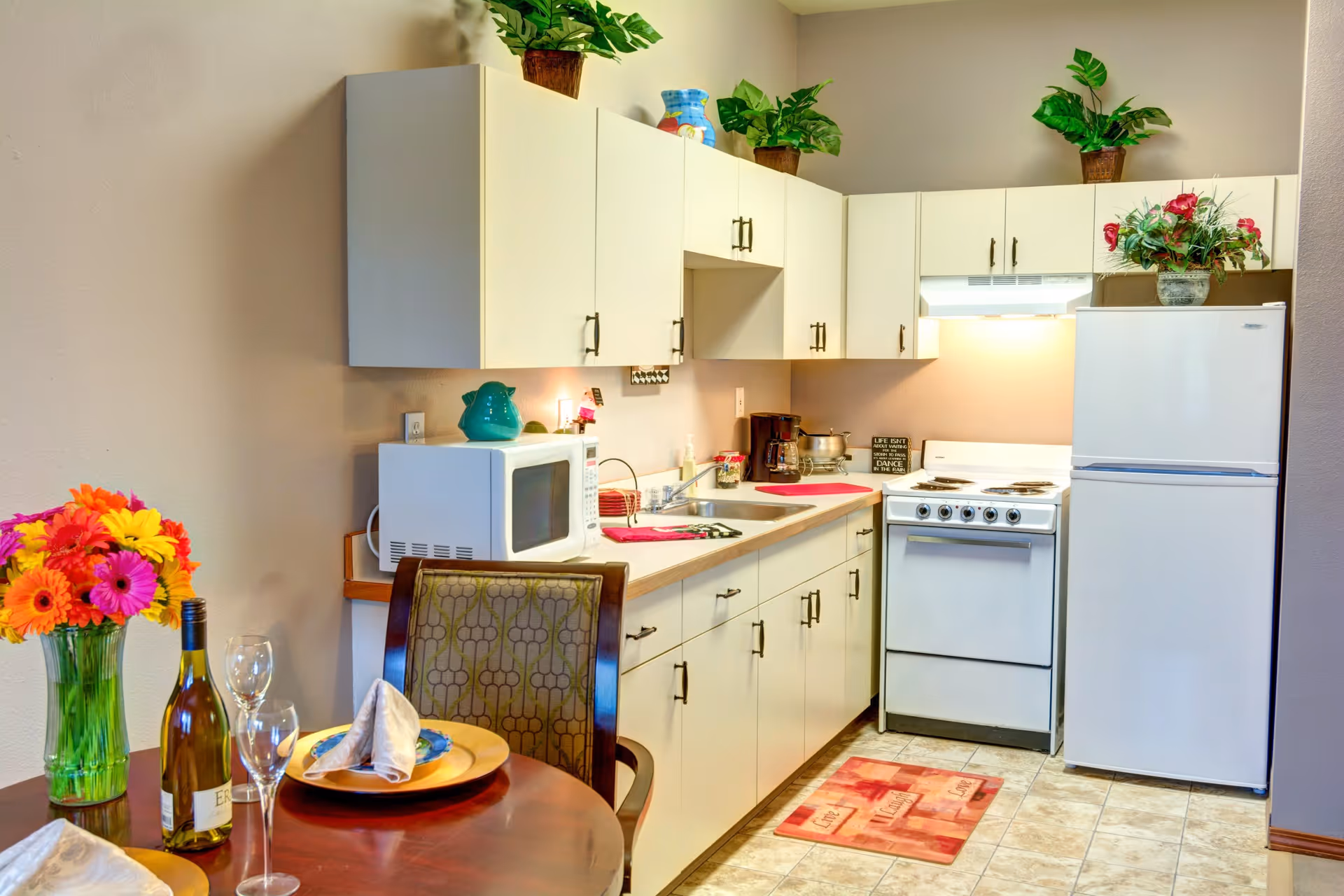 Compact kitchen and dining nook with white cabinets, a stove and refrigerator, a microwave on the counter, and a set table with flowers and wine.