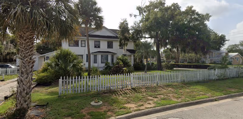 A two-story white house with a front porch surrounded by a white picket fence and lush greenery including palm trees and bushes, situated along a street with a grassy area in front.