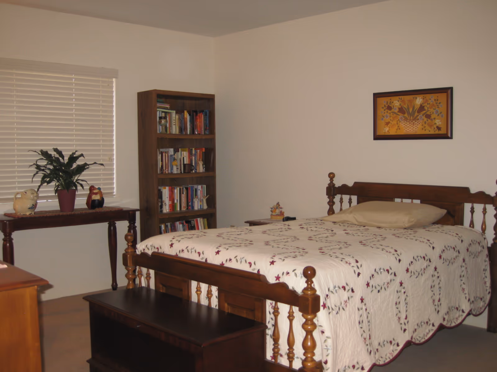 A bedroom with a wooden bed frame and a quilted bedspread featuring a floral pattern. There is a wooden bench at the foot of the bed, a wooden bookshelf filled with books, a table with decorative items including a potted plant, and a framed floral painting on the wall.