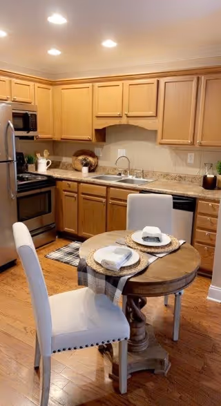 A cozy kitchen with wooden cabinets, granite countertops, and stainless steel appliances including a refrigerator, stove, microwave, and dishwasher. In the foreground, there is a small round wooden dining table set for two with white cushioned chairs and place settings with napkins on woven placemats. The floor is hardwood, and the kitchen is well-lit with recessed ceiling lights.