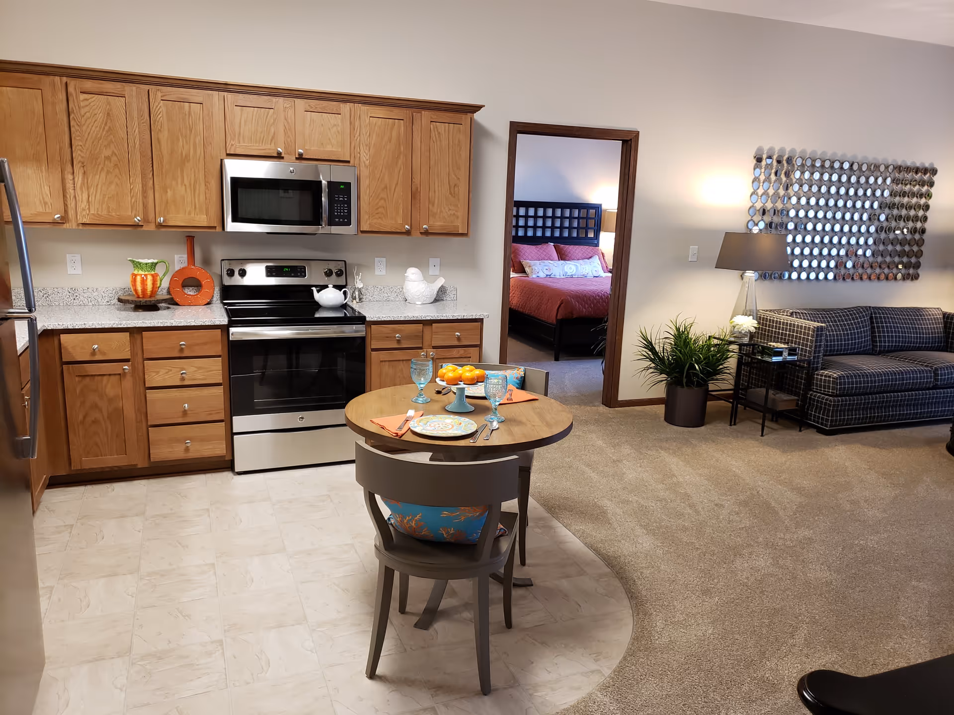 Interior view of a retirement community apartment showing a kitchen area with wooden cabinets, a stainless steel stove and microwave, and a small round dining table set for two. Adjacent to the kitchen is a living area with a checkered sofa, a side table with a lamp, a decorative wall mirror, and a potted plant. In the background, an open doorway reveals a bedroom with a bed dressed in red bedding.