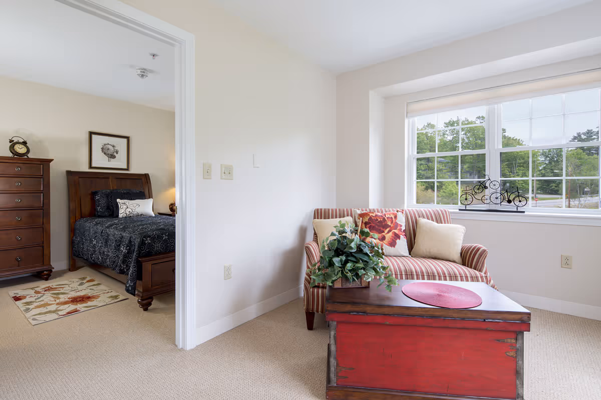 A cozy living area with a striped loveseat adorned with floral and beige pillows, a wooden coffee table with a red top and a potted plant, and a large window showing greenery outside. Adjacent to the living area is a bedroom with a wooden bed frame, black patterned bedding, a wooden dresser, a floral rug, and a framed flower picture on the wall.