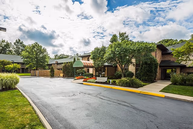 Exterior view of a senior living facility with a paved driveway, landscaped greenery, trees, and a building entrance with a green and orange striped awning under a partly cloudy sky.