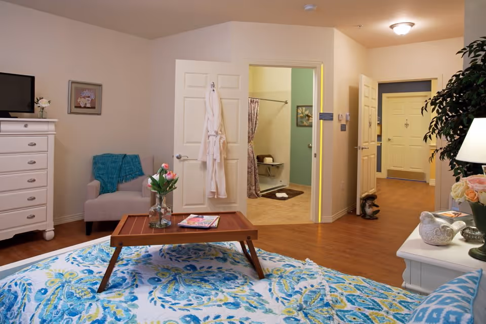 A cozy bedroom in a senior living facility with a bed covered in a blue and green patterned quilt. A wooden tray with a vase of flowers and magazines sits on the bed. To the left, there is a white dresser and a beige armchair with a teal throw blanket. The bathroom door is open, showing a walk-in shower with a curtain and a grab bar. The hallway and entrance door are visible in the background, with warm lighting and a potted plant on a white nightstand with a lamp and decorative items.