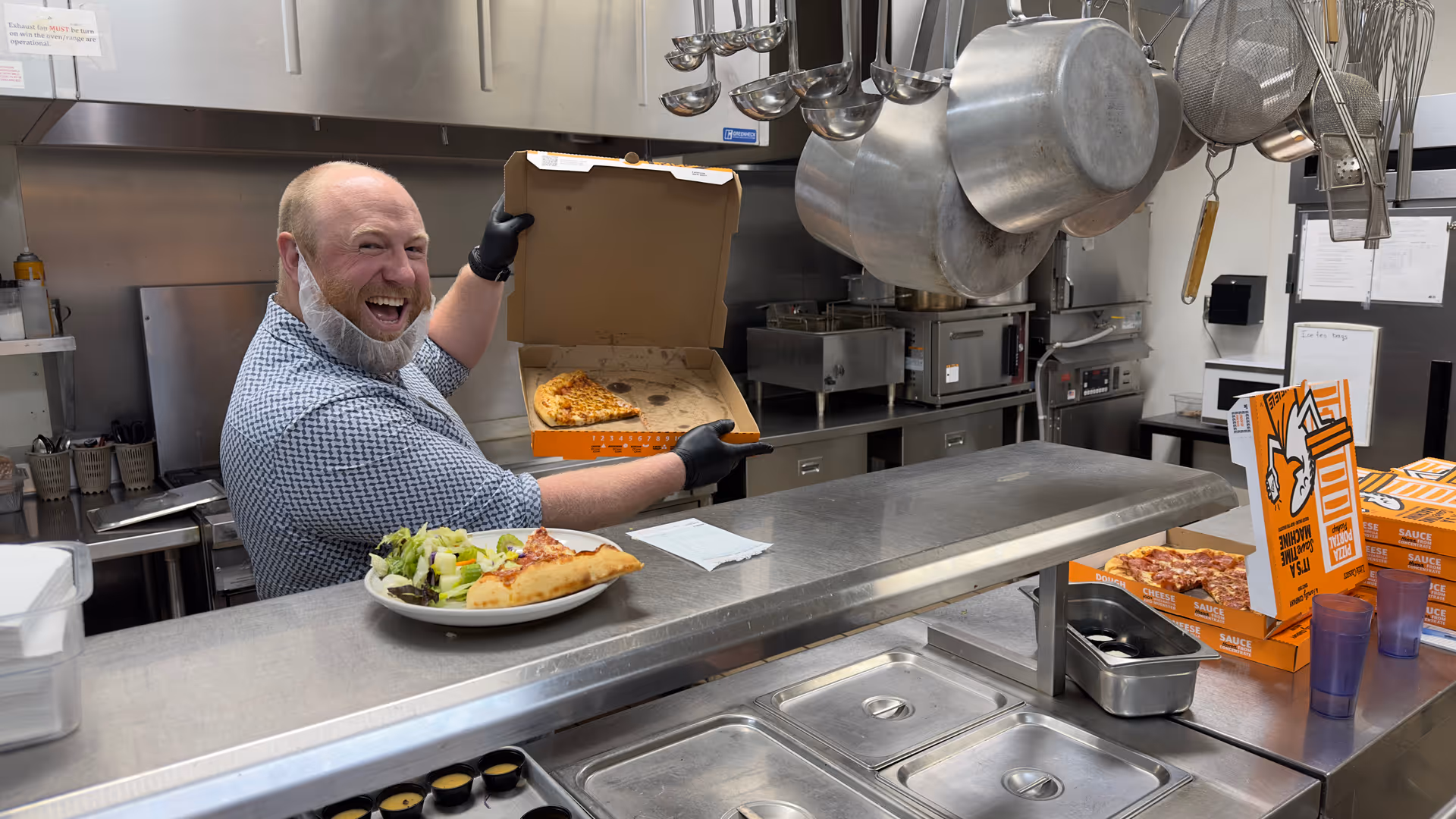 A man wearing a checkered shirt, black gloves, and a beard net is smiling and holding an open pizza box with a partially eaten pizza inside in a commercial kitchen. In front of him on the counter is a plate with a slice of pizza and a salad. The kitchen has stainless steel counters, hanging pots and ladles, and several pizza boxes stacked on the side.
