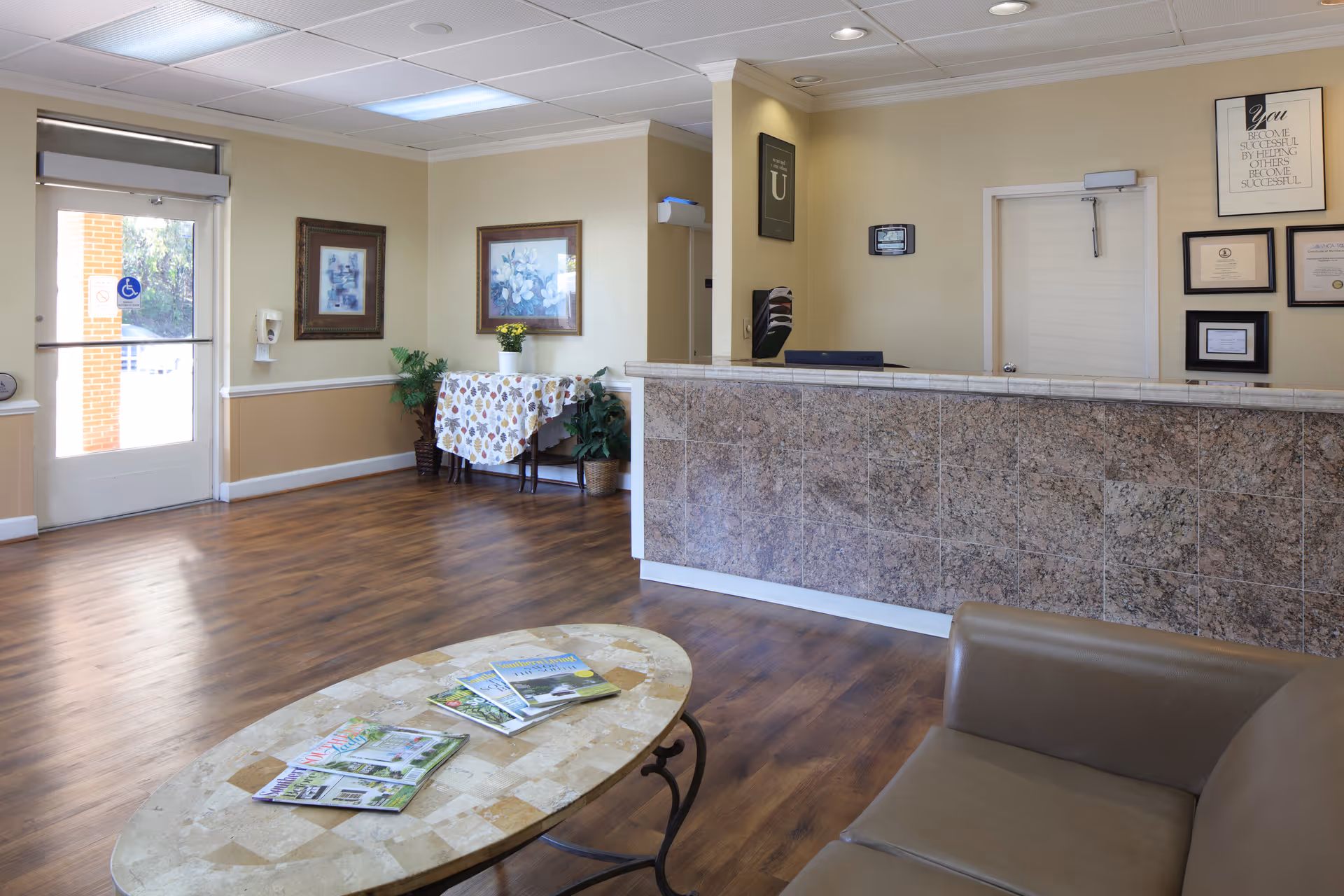 Reception area of Monroe Health and Rehabilitation Center with a marble front desk, a beige couch, a coffee table with magazines, framed pictures on the walls, a small table with a floral tablecloth and potted plants, and a door with a handicap accessibility sign.