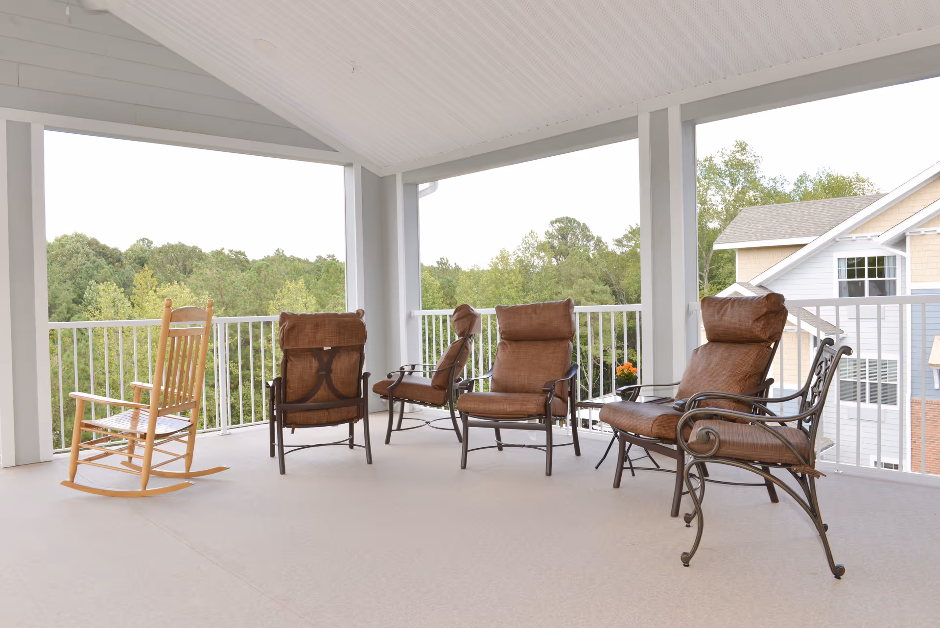 Covered outdoor balcony with cushioned metal armchairs and a wooden rocking chair overlooking trees and nearby buildings.