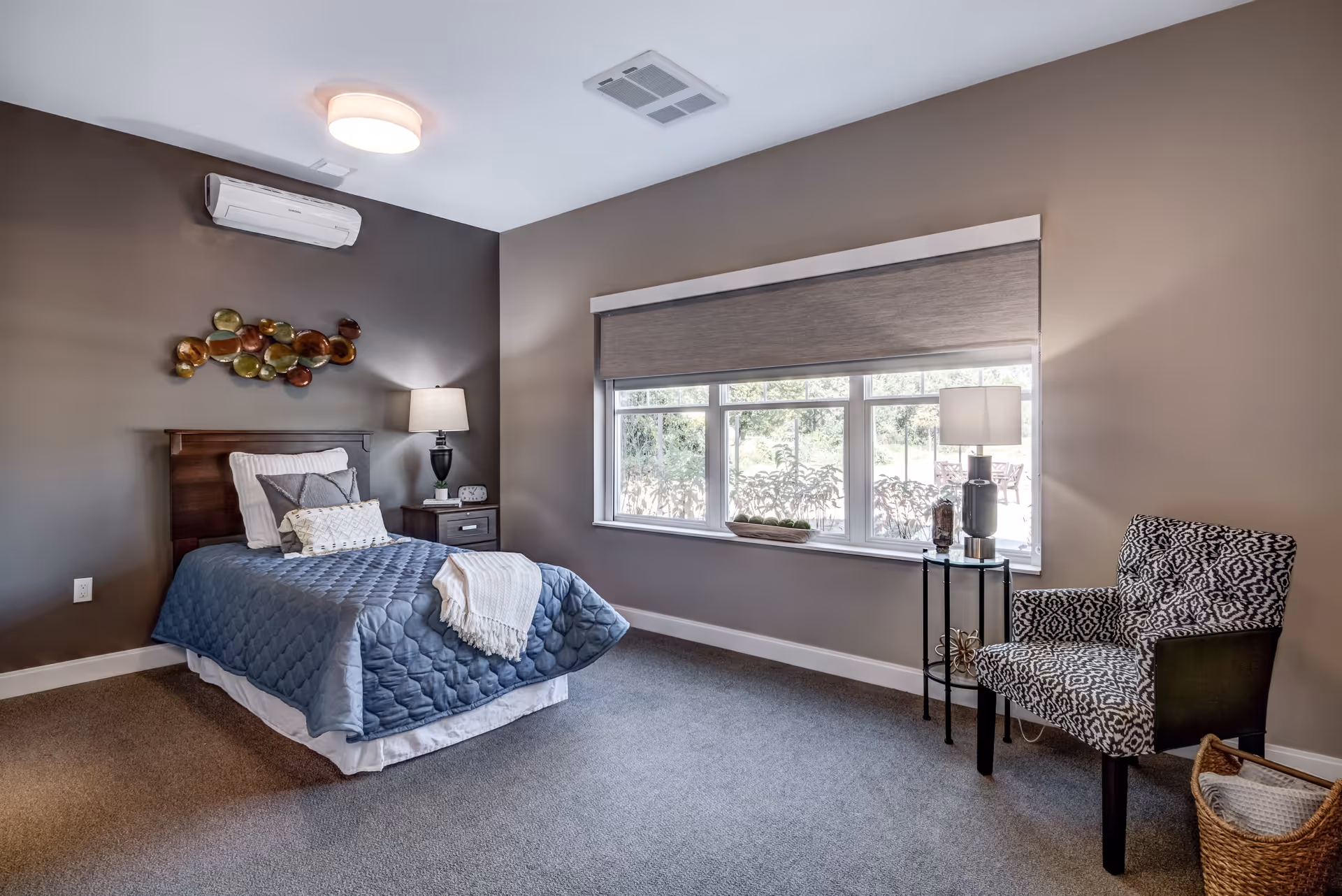 A neatly arranged bedroom with a single bed covered in a blue quilt and white pillows. There is a wooden headboard with a decorative wall art above it. A nightstand with a lamp and clock is beside the bed. A large window with a gray roller shade lets in natural light. Across from the bed is a patterned armchair next to a small round glass table with a lamp and decorative items. A wicker basket is on the floor near the chair.