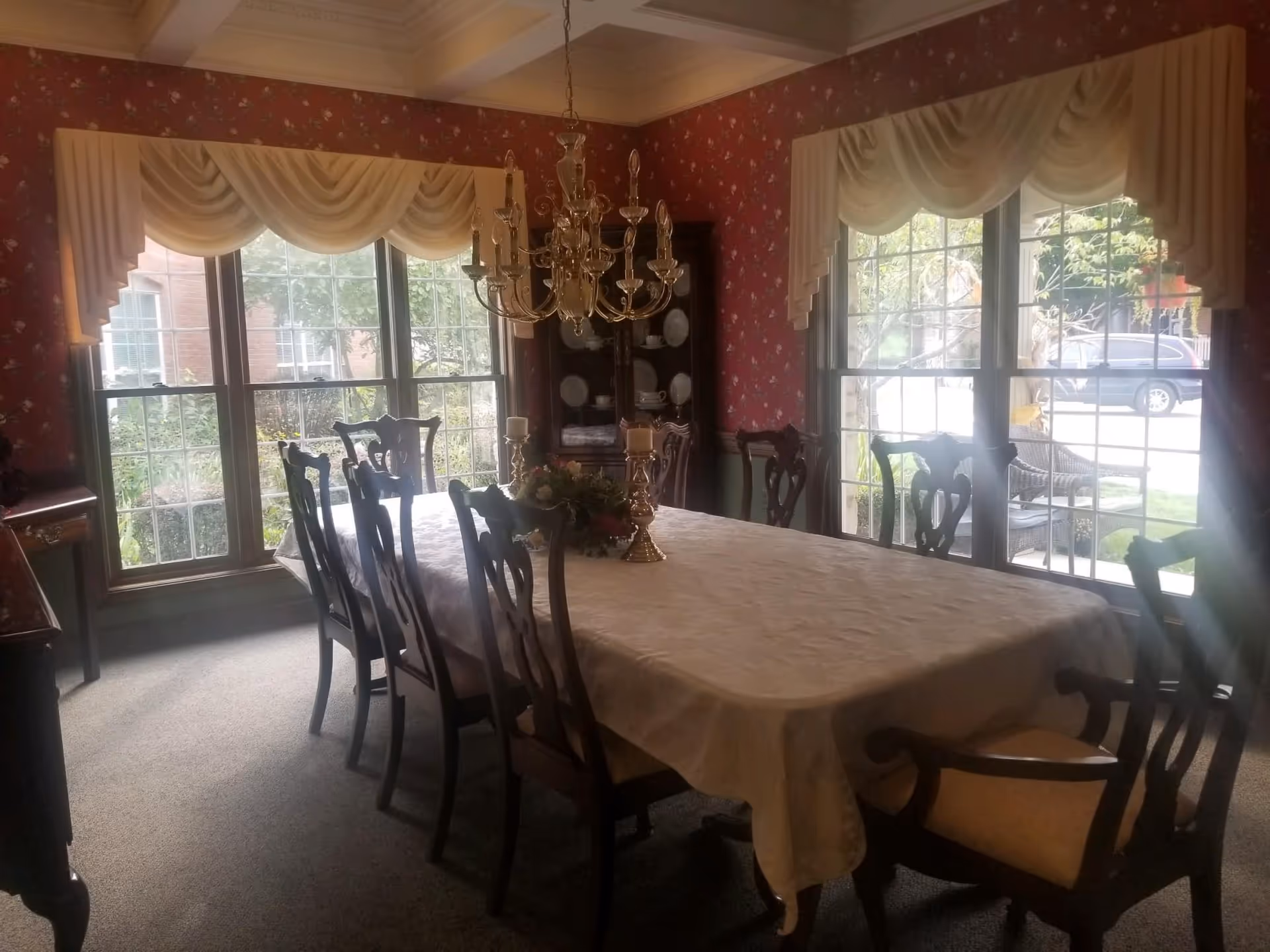 A traditional dining room with a long rectangular table covered with a white tablecloth, surrounded by wooden chairs with cushioned seats. The room has large windows with cream-colored valances, floral wallpaper in red tones, a chandelier hanging above the table, and a china cabinet displaying plates in the corner.