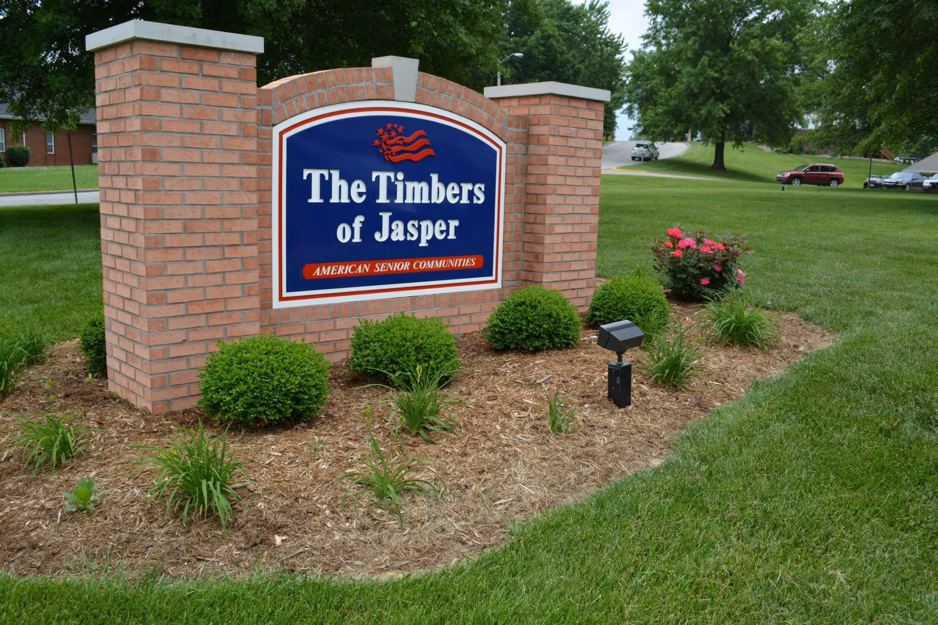 A brick sign for The Timbers of Jasper, an American senior community, surrounded by landscaped bushes and flowers with a grassy area and trees in the background.