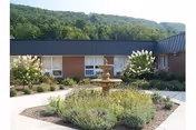 Outdoor courtyard area at Mountain View Rehabilitation and Senior Living Center featuring a central multi-tiered fountain surrounded by landscaped bushes and flowering plants, with a building and forested hills in the background under a clear sky.
