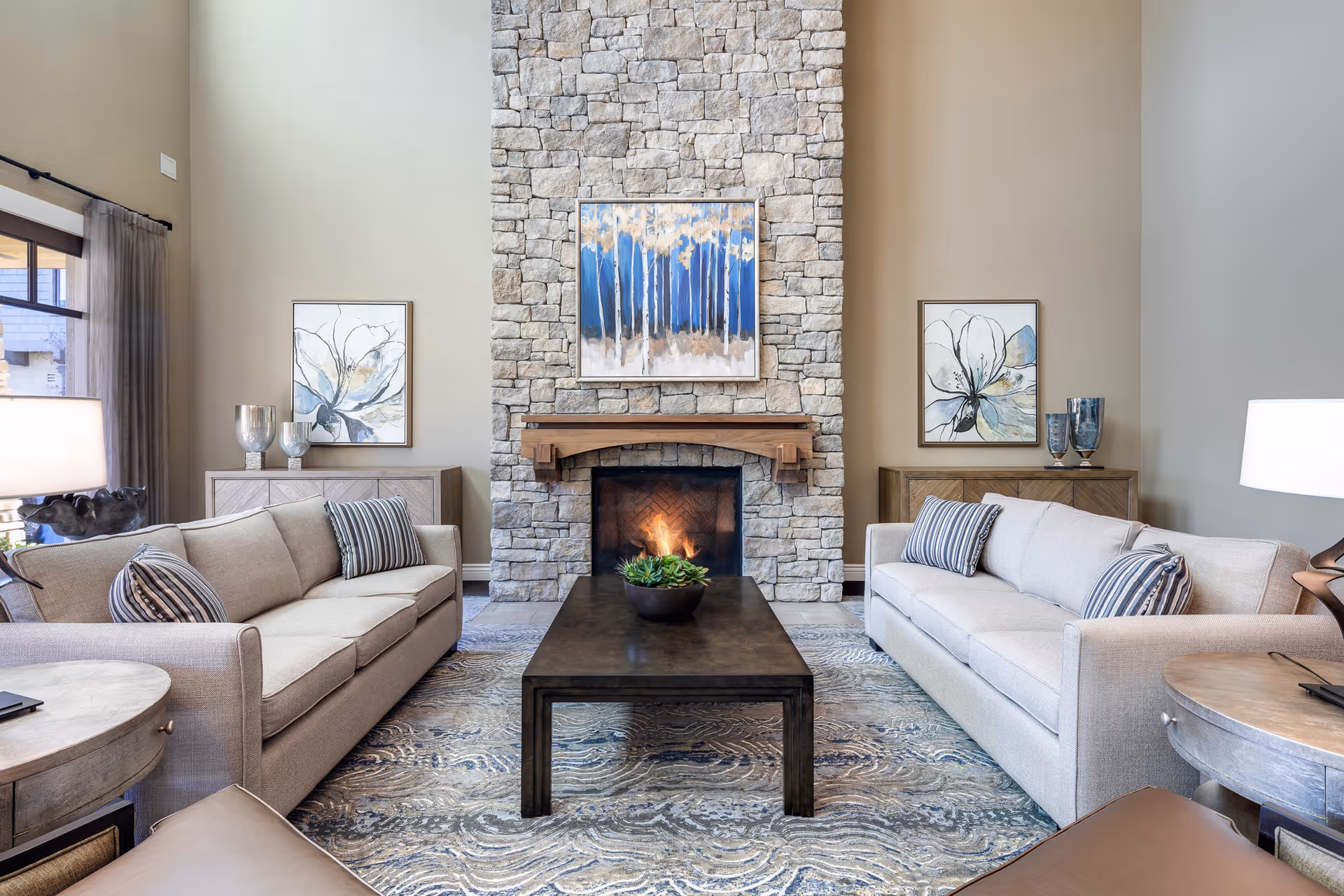 A cozy living room with two beige sofas facing each other across a dark wooden coffee table. The room features a tall stone fireplace with a fire burning inside, flanked by two side tables with decorative vases and framed floral artwork on the walls. The space is well-lit with natural light from a window on the left and table lamps on either side.