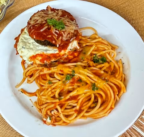 A white plate with a serving of spaghetti topped with tomato sauce and melted cheese, accompanied by a portion of eggplant parmesan garnished with shredded cheese and a small sprig of parsley.