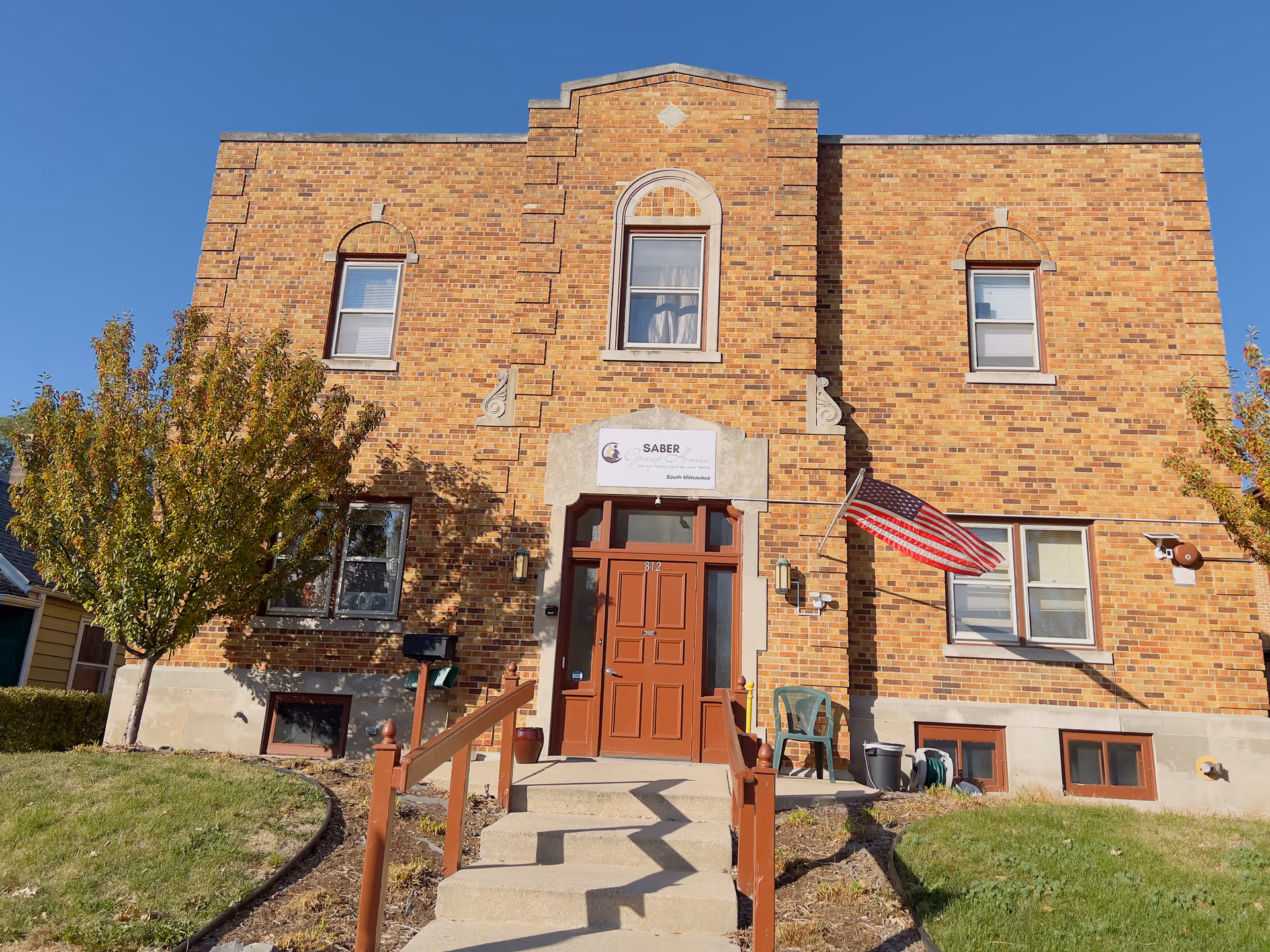 Front exterior view of a two-story brick building with a central entrance featuring a brown door and steps leading up to it. There are windows on both floors, an American flag mounted on the right side, and two small trees flanking the entrance. A sign above the door reads 'SABER Group Homes South Milwaukee.'