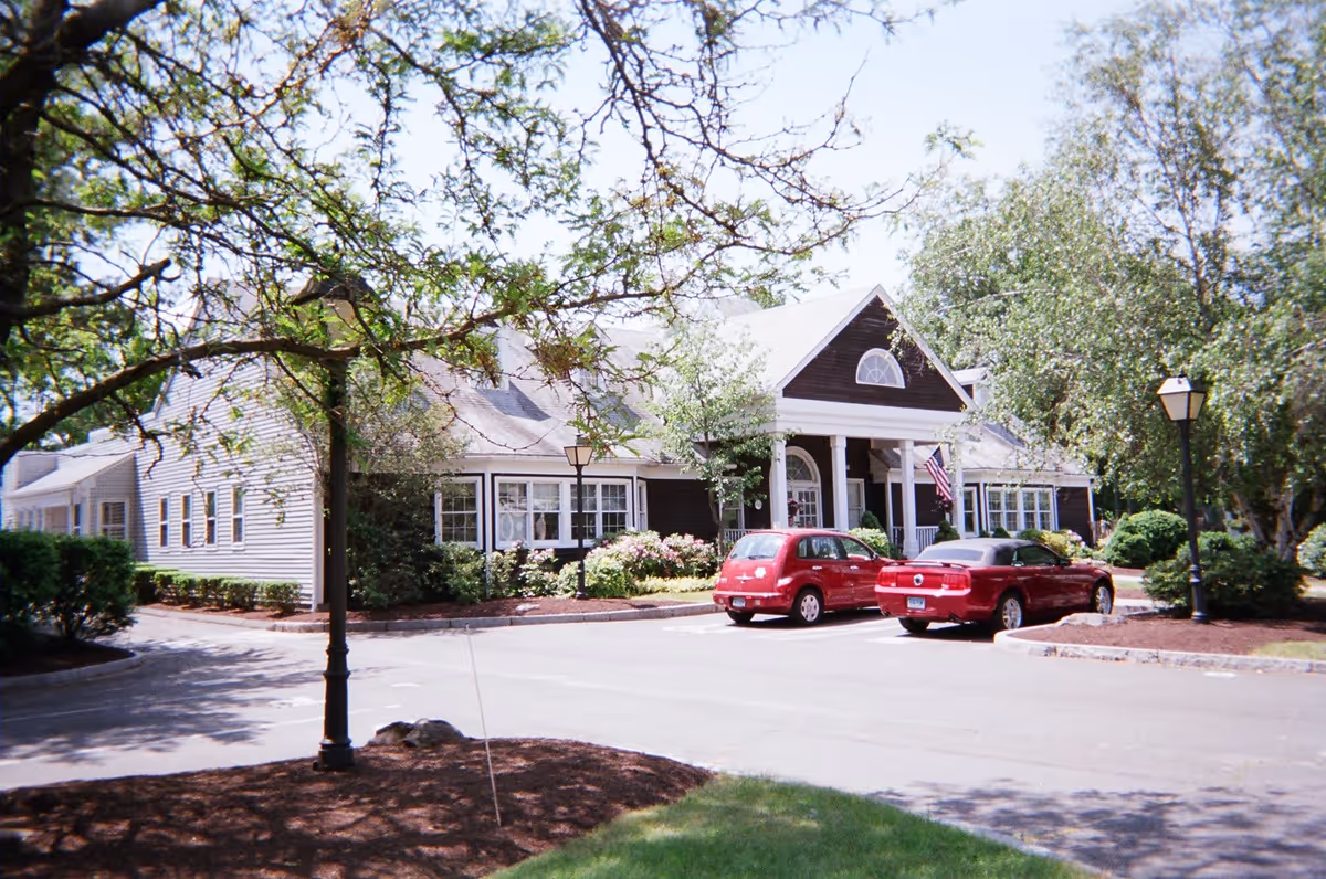 Exterior view of a single-story building with a gabled roof, surrounded by trees and shrubs. Two red cars are parked in front of the building near a driveway. The building has white columns at the entrance and an American flag displayed near the door.