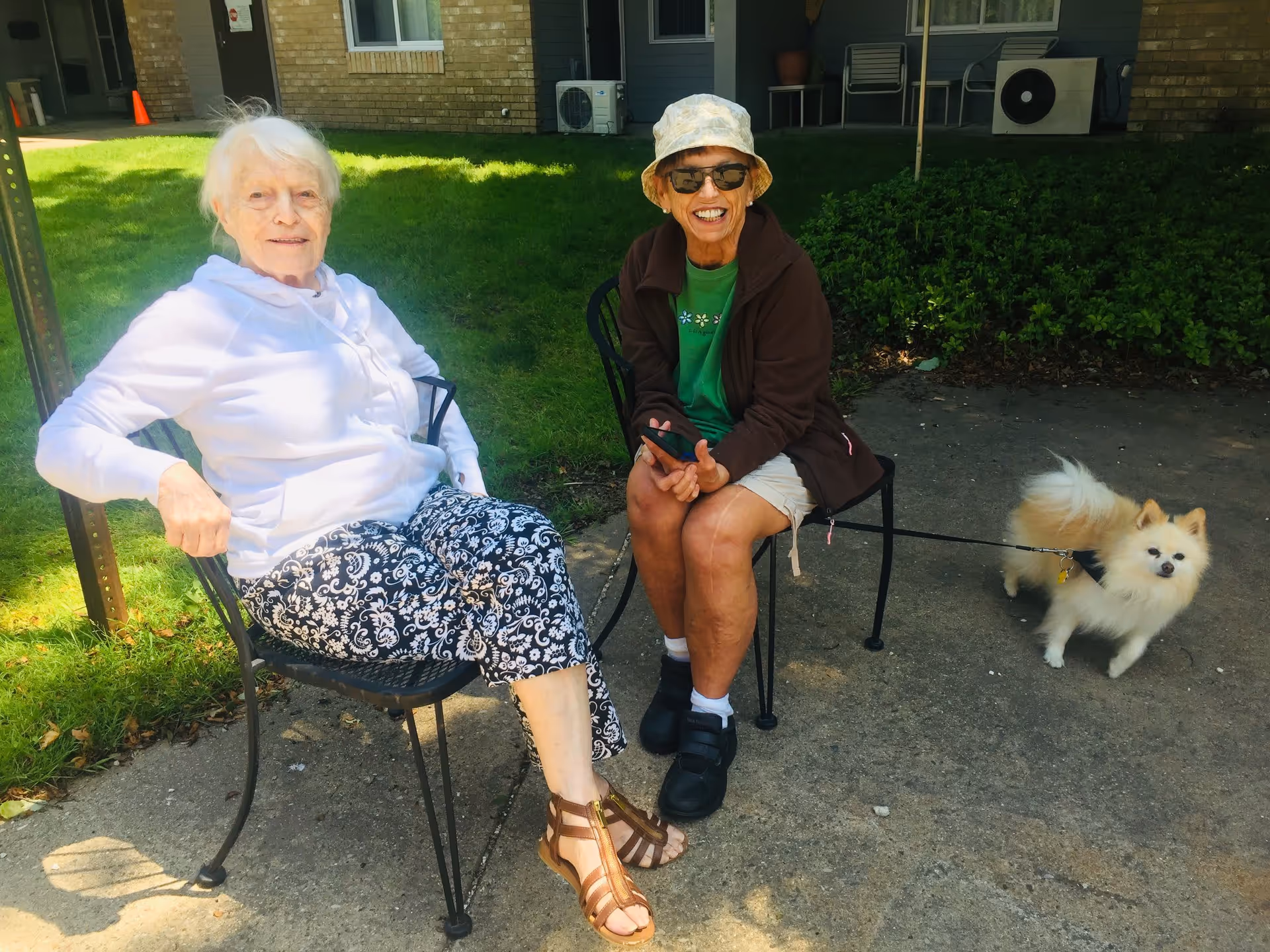 Two elderly women sitting on metal chairs outside on a sunny day. One woman is wearing a white hoodie and patterned pants, while the other is wearing a green shirt, brown jacket, shorts, sunglasses, and a hat. A small fluffy dog on a leash is standing nearby on the pavement. Behind them is a building with windows and some greenery.