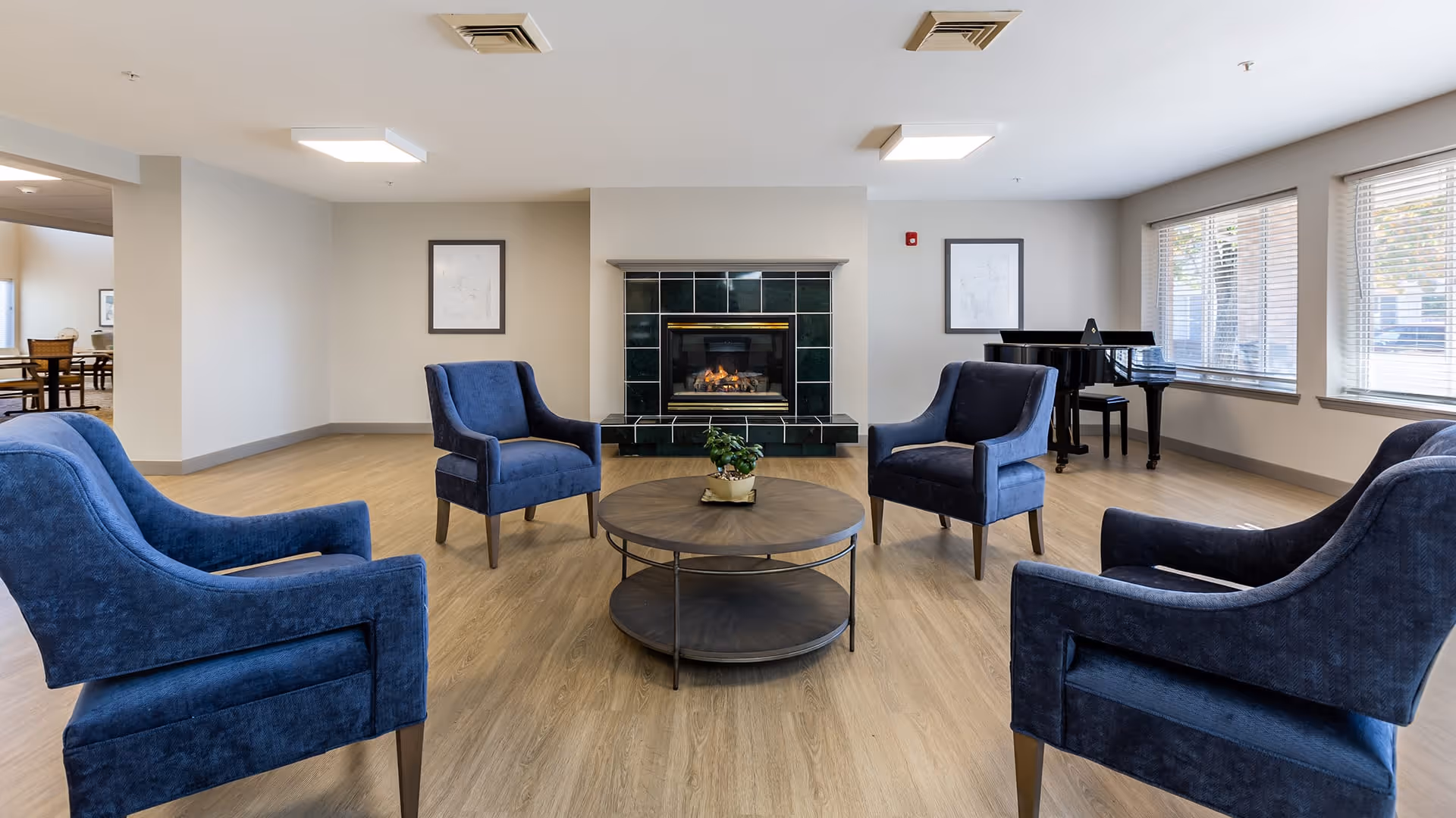 A bright and spacious living room area with four blue upholstered armchairs arranged around a round wooden coffee table. A fireplace with a black tile surround is centered on the far wall, flanked by two framed pictures. Large windows on the right side let in natural light, and a black grand piano is positioned near the windows. The room has light wood flooring and white walls.