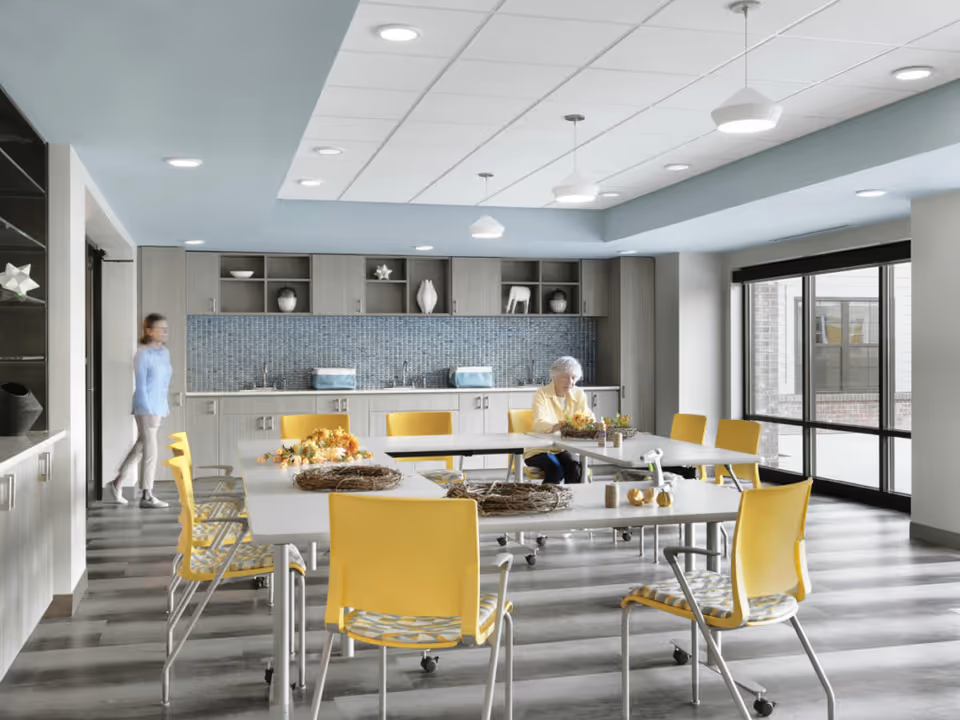 A bright and modern activity room in a senior living facility with large windows letting in natural light. The room features several tables arranged in a U-shape with yellow chairs around them. An elderly woman is seated at one of the tables working on a craft project with autumn-themed decorations. In the background, there is a kitchenette area with cabinets, a blue tiled backsplash, and decorative items on the shelves. A younger person is walking in the background near the kitchenette.