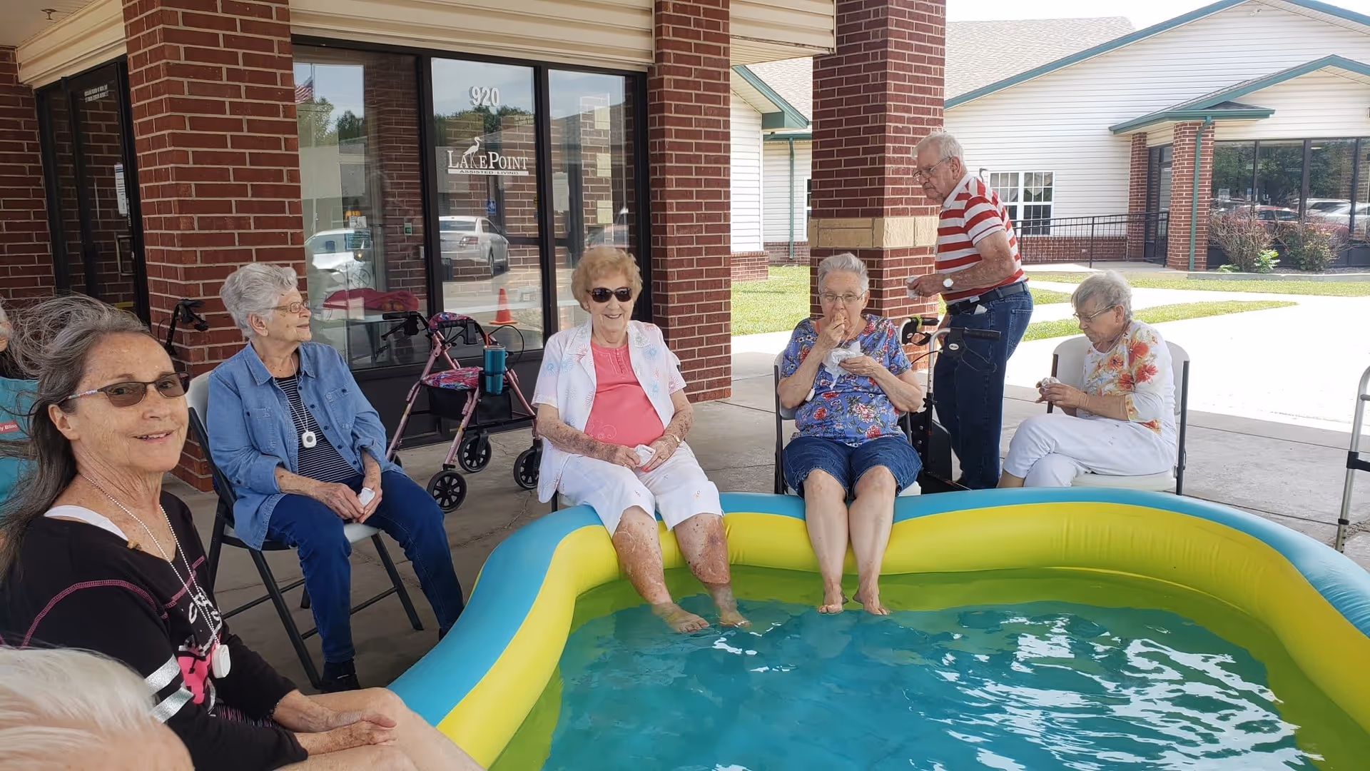 A group of elderly people sitting around a small inflatable pool filled with water outside a brick building. Some have their feet in the pool, and one person is standing nearby. The building has a sign that reads 'LakePoint Augusta'.