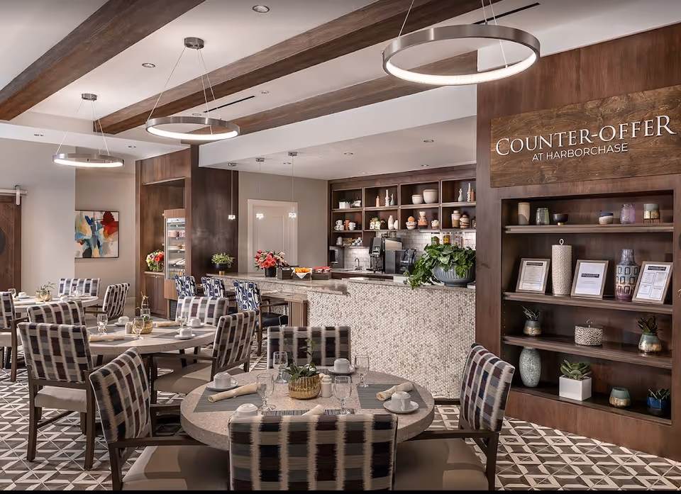 Modern dining room with round tables and patterned chairs facing a service counter and shelving labeled Counter-Offer at Harborchase.