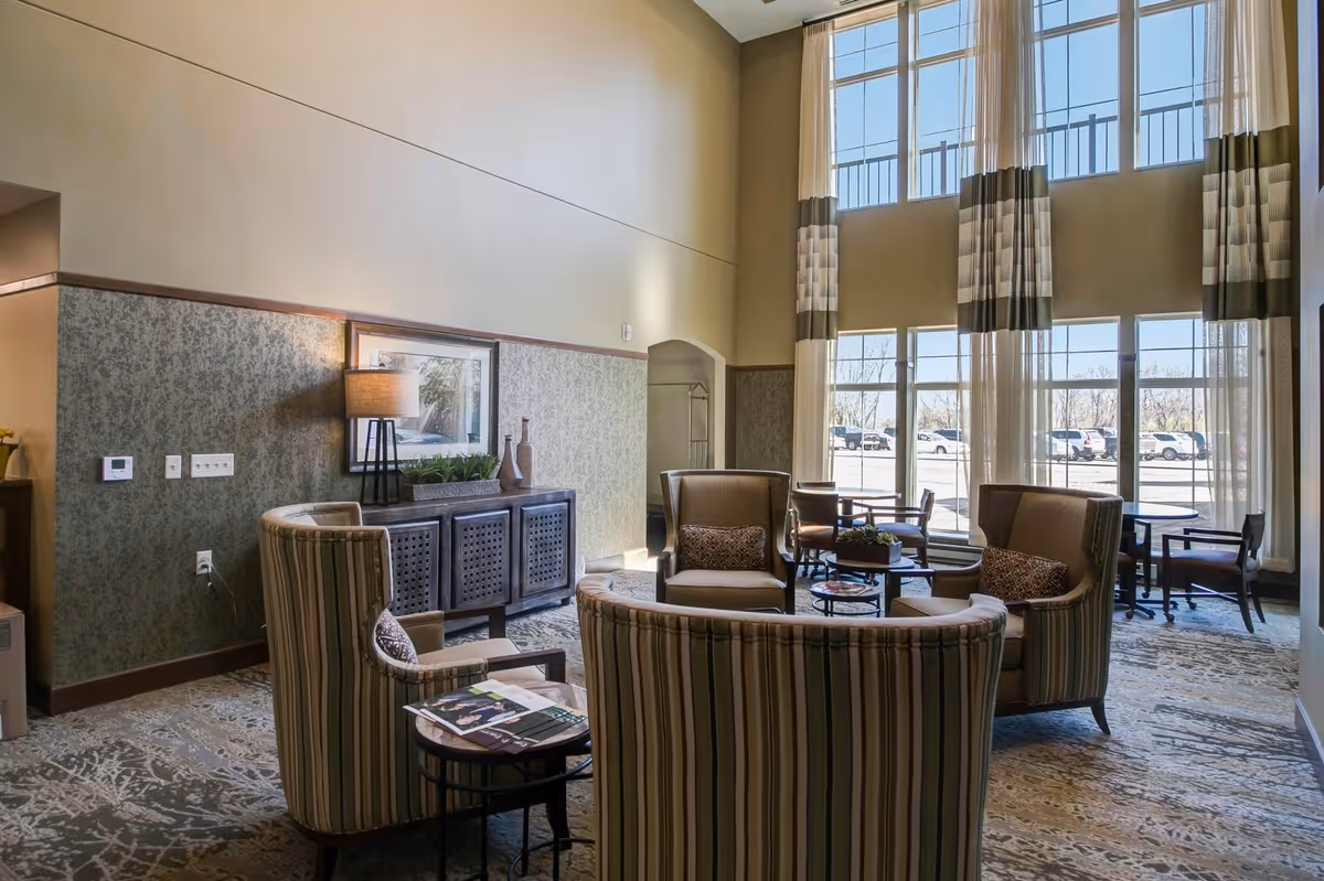 A bright and spacious common area with high ceilings and large windows letting in natural light. The room features a seating arrangement with striped upholstered chairs and a curved sofa around a small table. There is a sideboard with a lamp, decorative vases, and a framed picture on the wall. Sheer curtains hang from the tall windows, and outside, parked cars are visible.