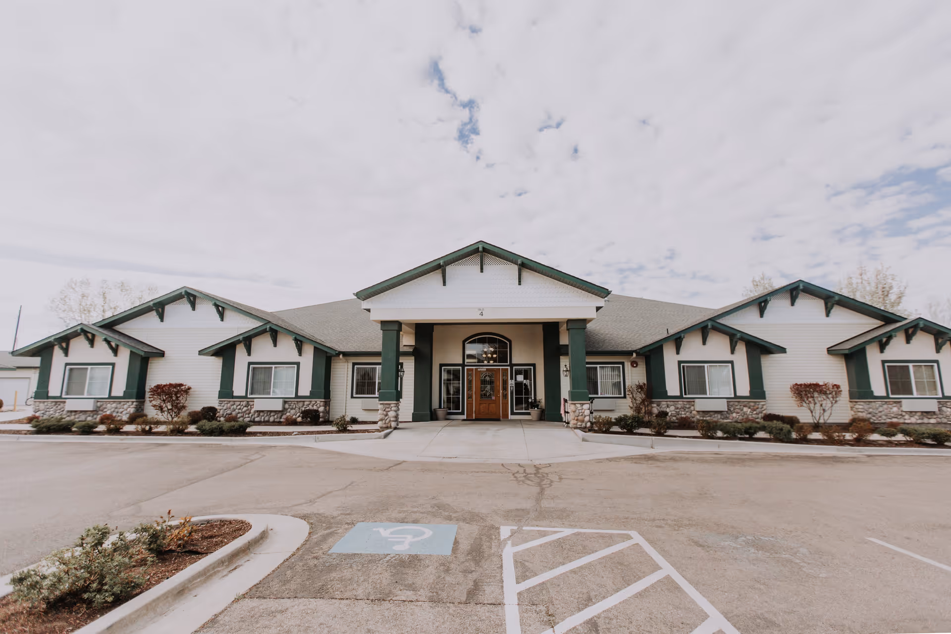 Front exterior view of a single-story assisted living facility building with white siding, green trim, and stone accents. The entrance has a covered porch supported by green columns, and there is a handicapped parking space visible in the foreground.