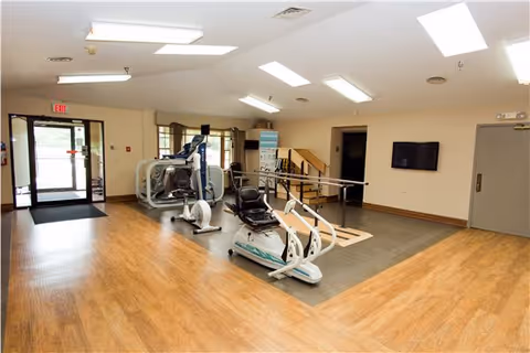 Interior view of a senior living facility exercise room with wooden flooring and exercise equipment including a recumbent bike, rowing machine, and parallel bars. There is a TV mounted on the wall, a door, and an exit door with windows allowing natural light to enter the room.
