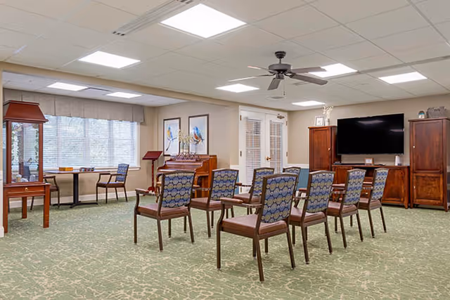 A senior living activity room with rows of chairs facing a television and piano, wood cabinets, and large windows.