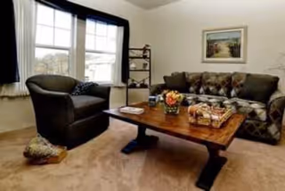 A cozy living room with a large window covered by white curtains and dark valances, a dark upholstered armchair, a patterned sofa, and a wooden coffee table with decorative items including a flower arrangement and a basket. A framed picture hangs on the wall above the sofa.