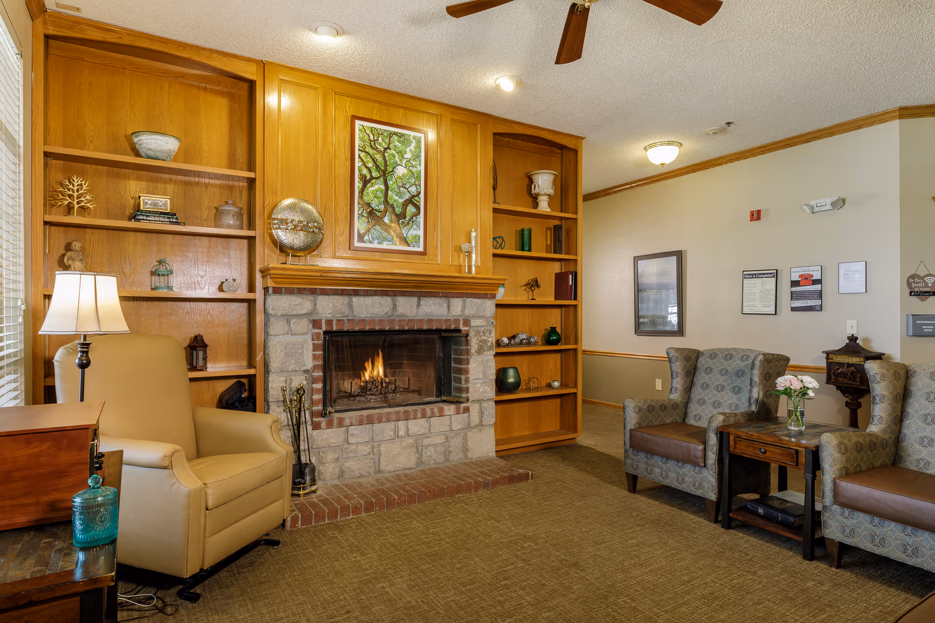 A cozy living room area with a stone fireplace in the center, wooden built-in shelves on either side filled with decorative items, a beige armchair with a floor lamp beside it on the left, and two patterned armchairs with a small wooden table holding a vase of flowers on the right. The walls are light-colored with wood trim, and there are framed pictures and notices on the far wall.
