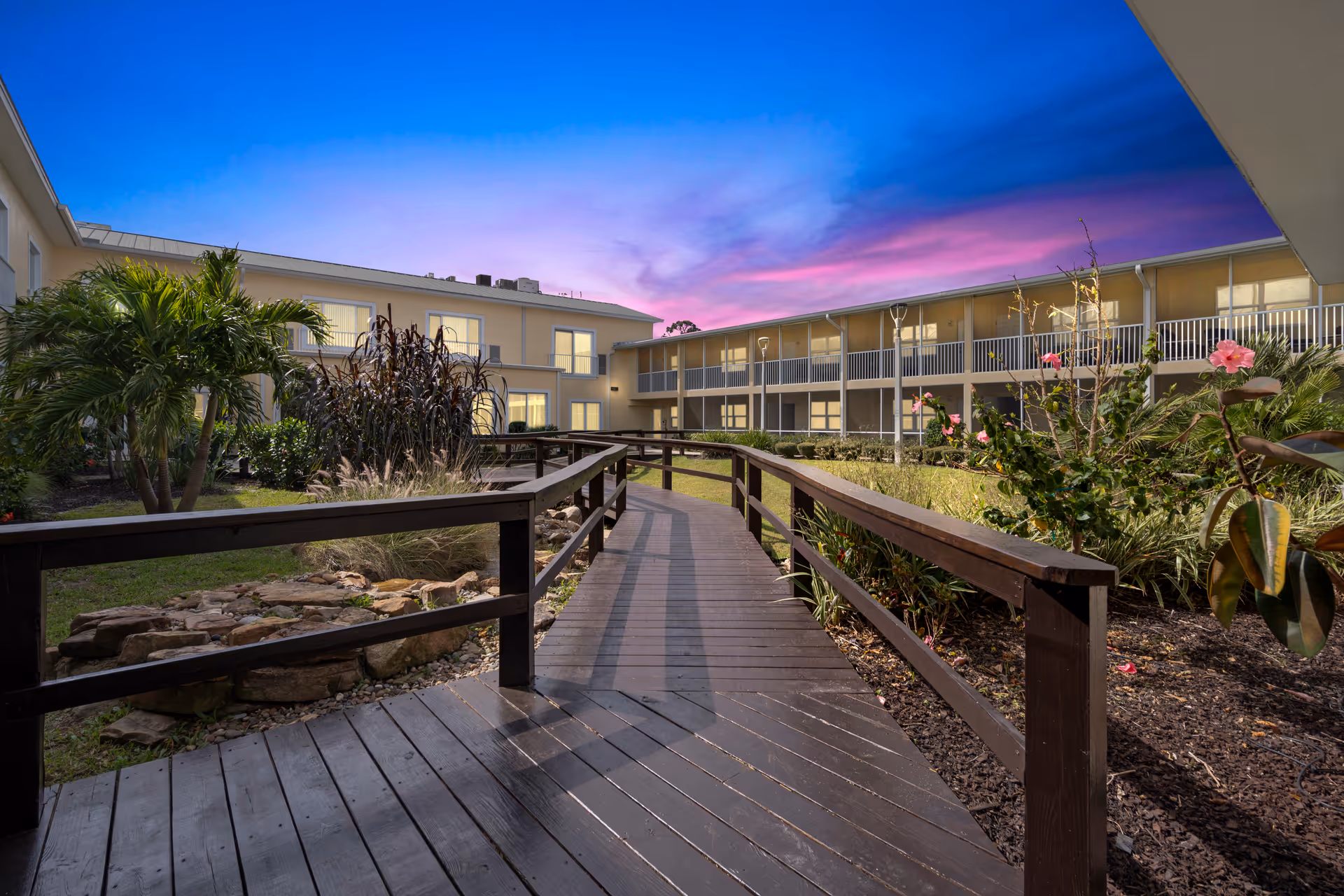Outdoor view of a senior living facility courtyard at dusk with a wooden walkway surrounded by landscaped plants and flowers, and two-story buildings with balconies and windows in the background under a colorful sky.