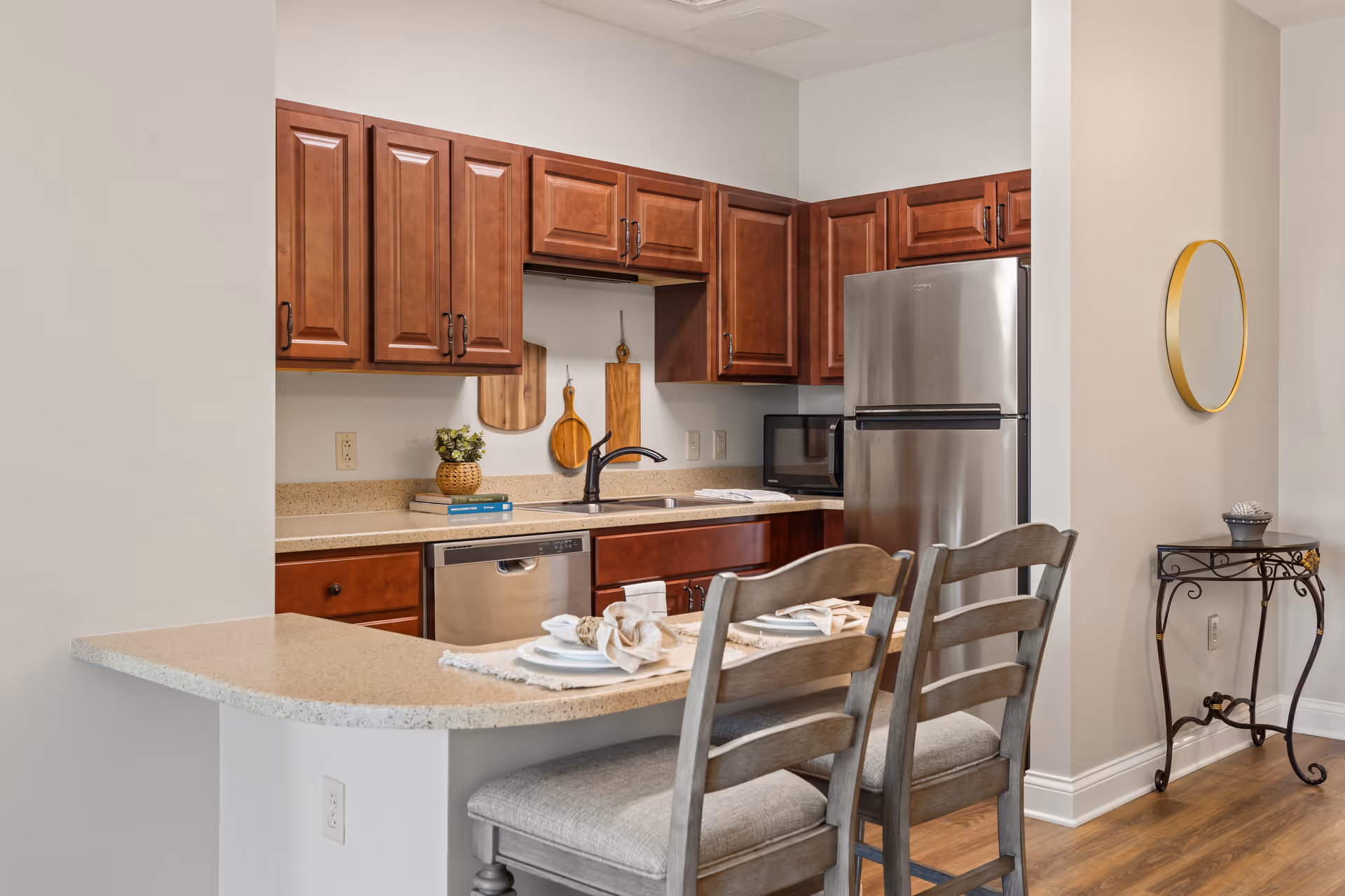A modern kitchen with wooden cabinets, a stainless steel refrigerator, dishwasher, and microwave. The countertop extends into a breakfast bar with two cushioned chairs set with plates and napkins. A small decorative table with a round mirror hangs on the adjacent wall.