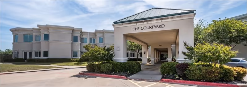 Exterior view of a senior living facility named The Courtyard, showing a covered entrance with columns, surrounded by greenery and a clear blue sky.