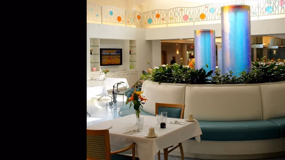 A bright and modern dining area in a senior living facility with a table set for two, featuring white tablecloth, glassware, and a vase with orange flowers. In the background, there is a circular seating area with green plants and two tall, illuminated blue cylindrical decorations. A television is mounted on the far wall, and a walker is visible near the seating area.