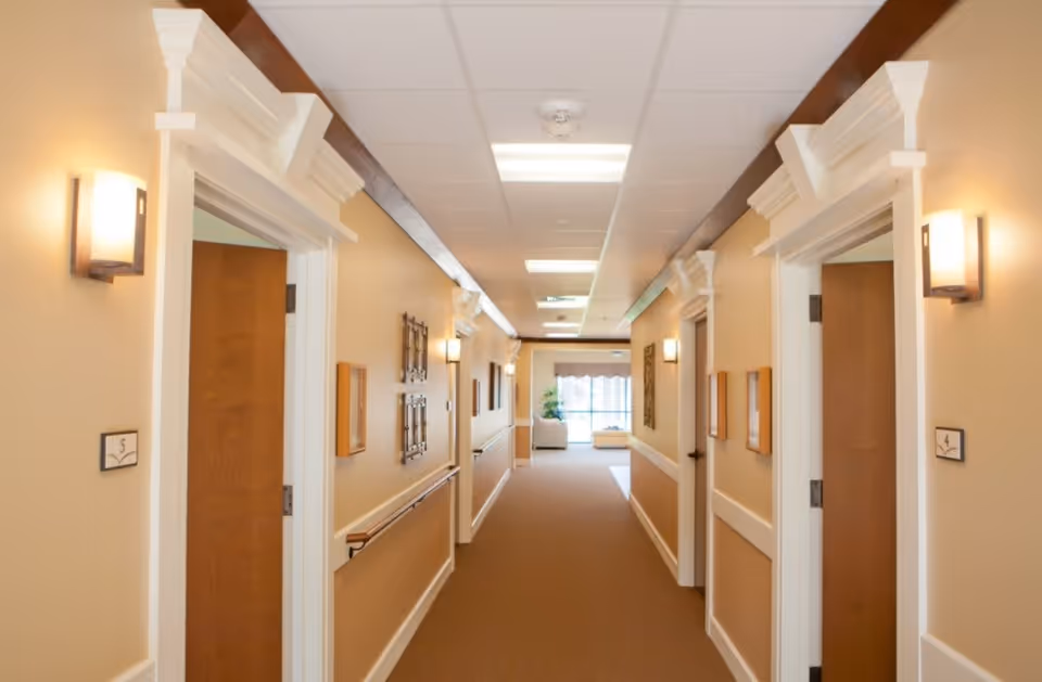 A well-lit hallway in a senior living facility with beige walls, white trim, and brown carpet. Doors to rooms are visible on both sides, each with a small sign indicating room numbers. Handrails run along the walls, and decorative wall sconces provide lighting. At the end of the hallway, there is a seating area with a window letting in natural light.