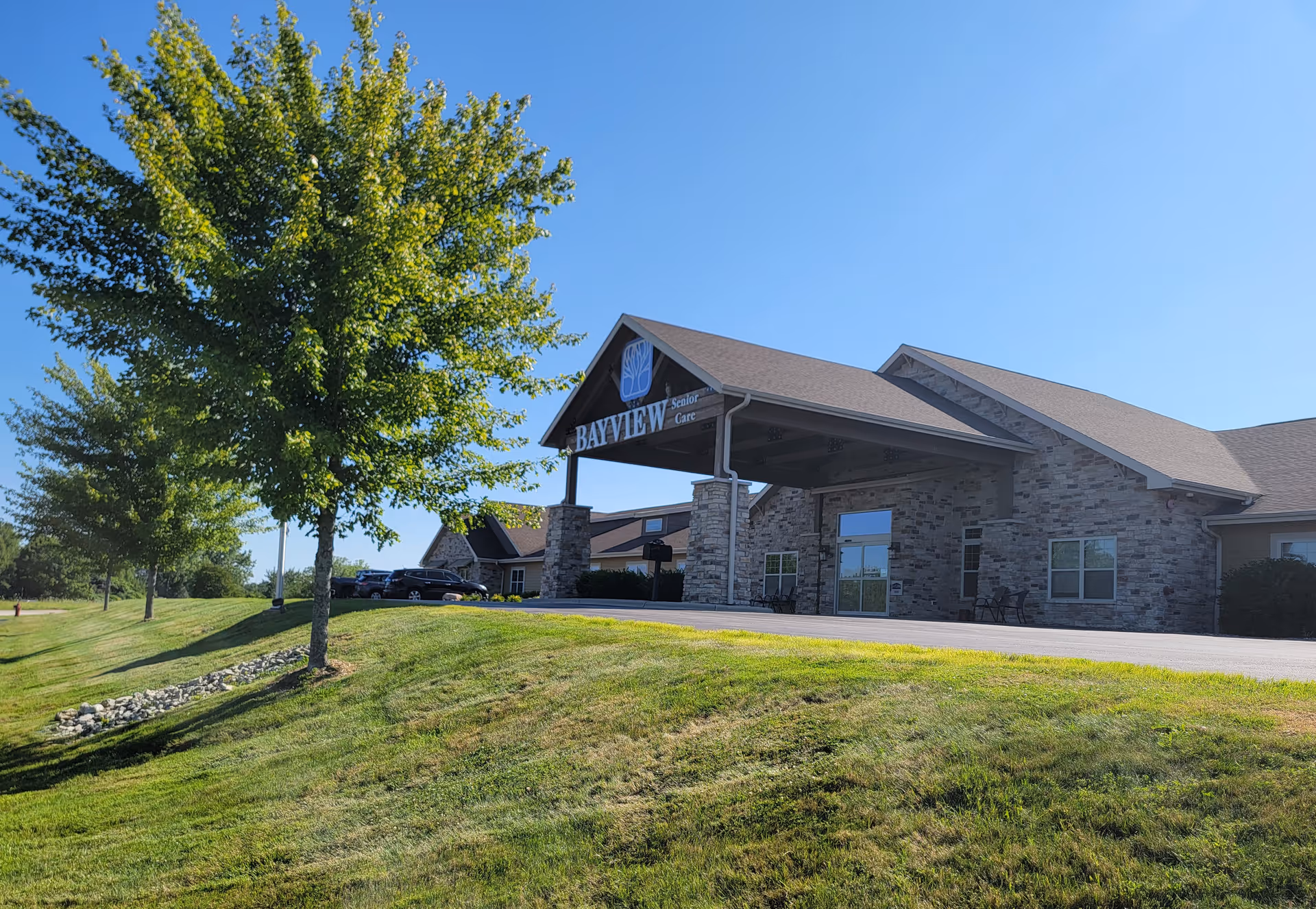 Exterior view of Bayview Senior Care building with stone facade, a covered entrance supported by stone pillars, and a well-maintained grassy area with trees under a clear blue sky.