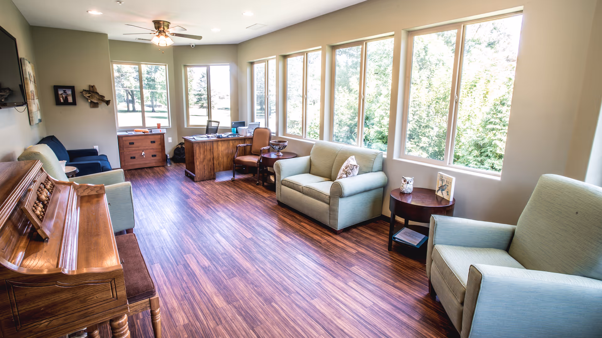 Bright common lounge with a piano, multiple armchairs and sofas, a desk, and large windows overlooking trees.