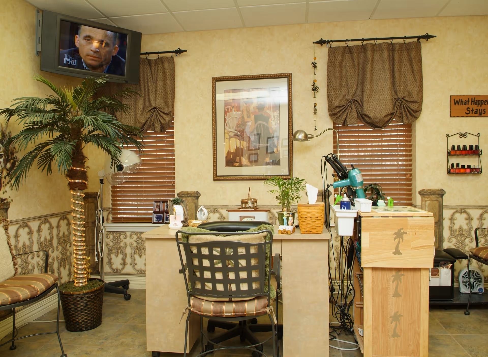 Interior of a salon area in a senior living community with a hair styling station, chairs, a potted palm tree, two windows with brown curtains, a wall-mounted TV showing Dr. Phil, and various salon equipment including a hair dryer and nail polish rack.