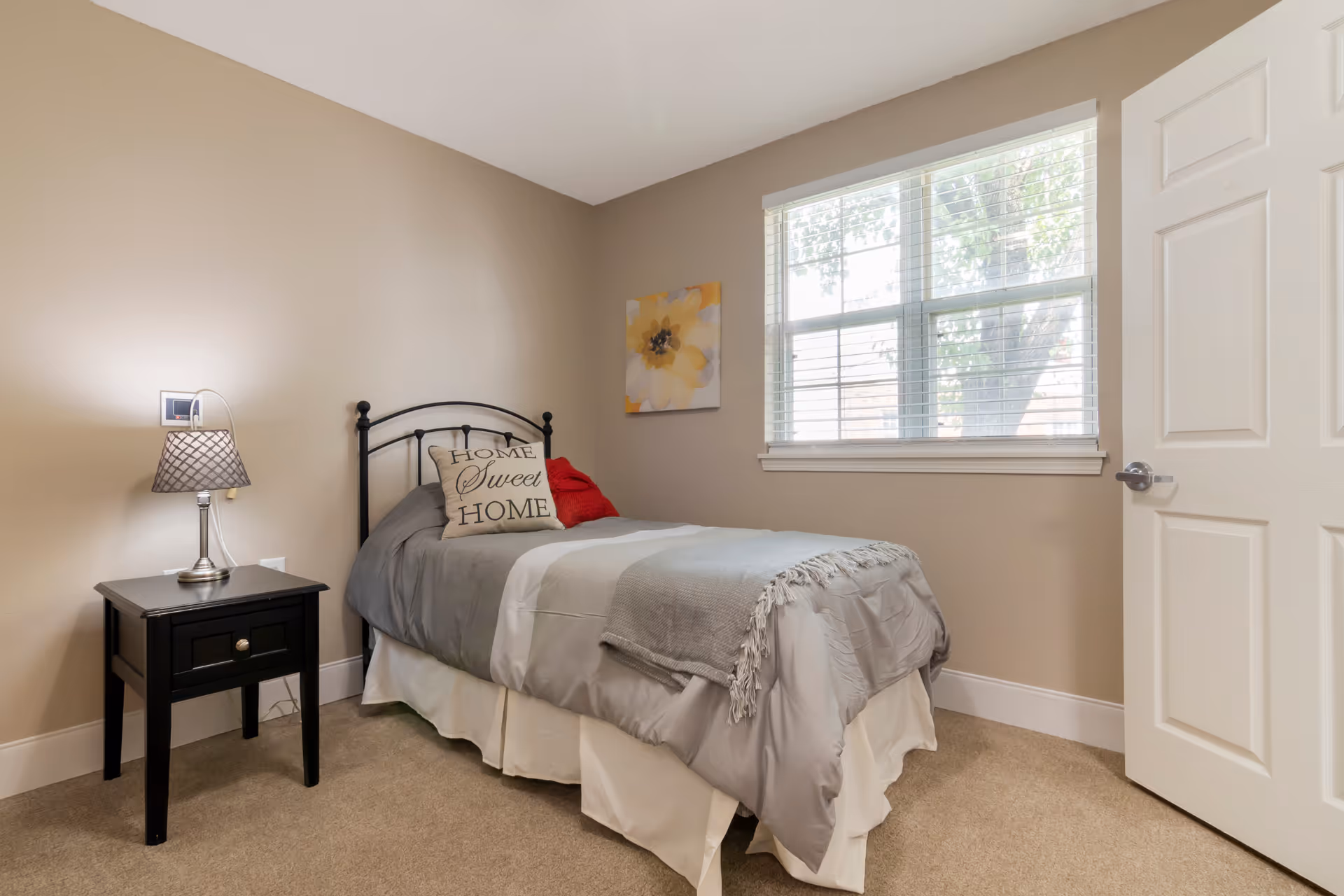 A small bedroom with beige walls and carpet, featuring a single bed with gray and white bedding, a pillow that says 'Home Sweet Home,' and a red pillow. Next to the bed is a black nightstand with a lamp. A window with white blinds lets in natural light, and a white door is partially open.