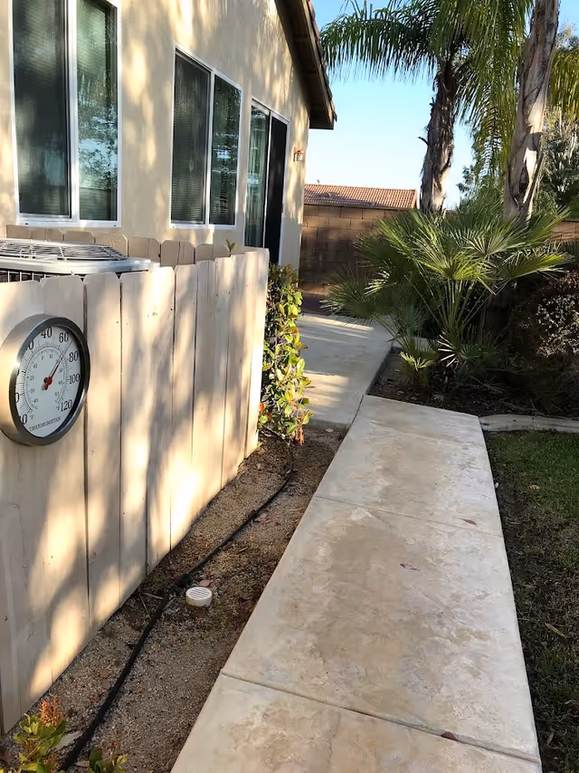 Concrete walkway along the side of a house with a white fence-mounted thermometer, windows, and palm trees in the yard.