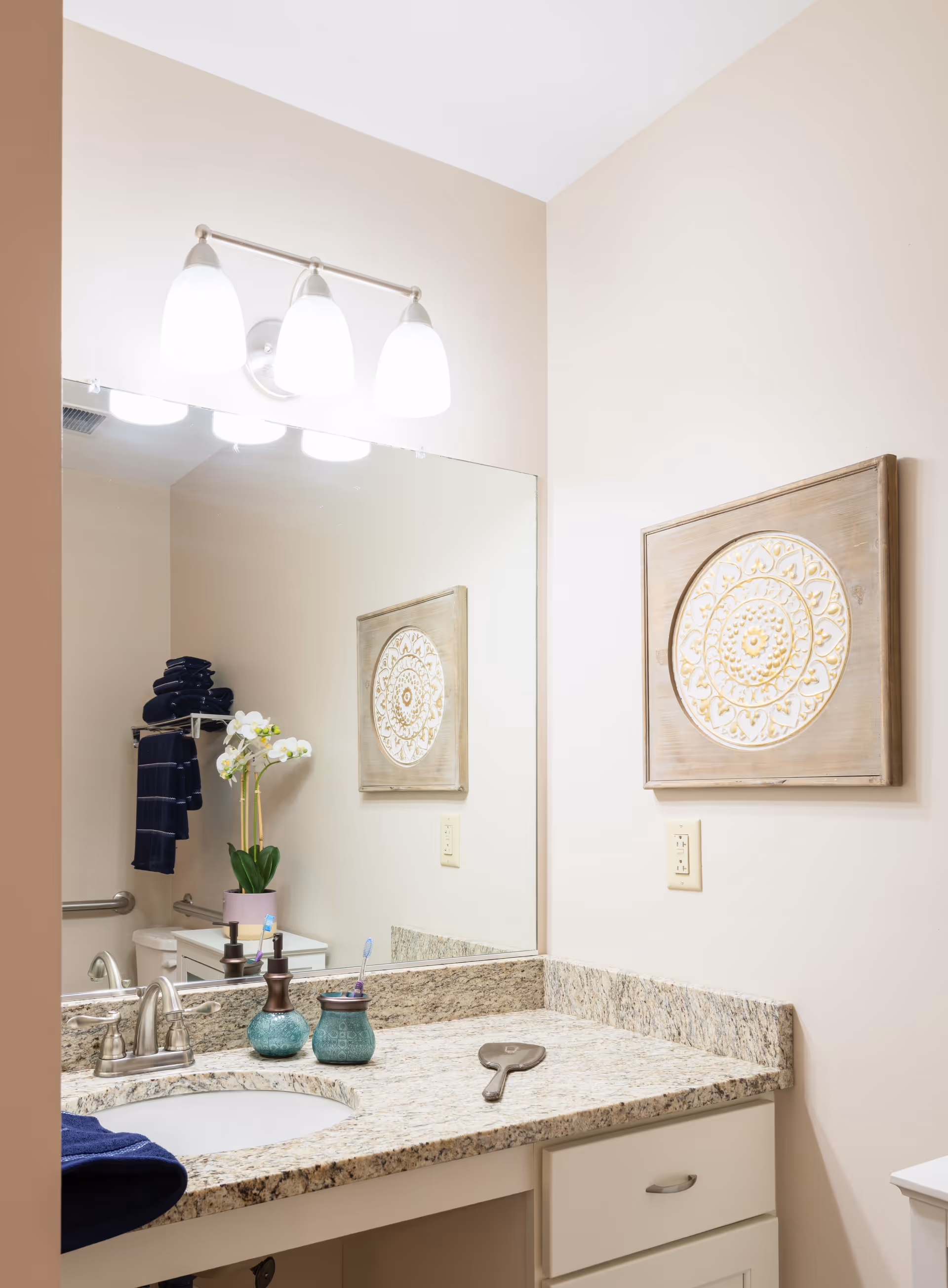 A clean and well-lit bathroom vanity area with a granite countertop, an under-mount sink, and a silver faucet. On the countertop are two decorative containers, a hand mirror, and a toothbrush. A large mirror is mounted on the wall above the sink, reflecting a towel rack with dark blue towels and a white orchid plant. Above the mirror is a three-light fixture with frosted glass shades. A decorative wooden wall art piece with a circular carved design hangs on the adjacent wall.