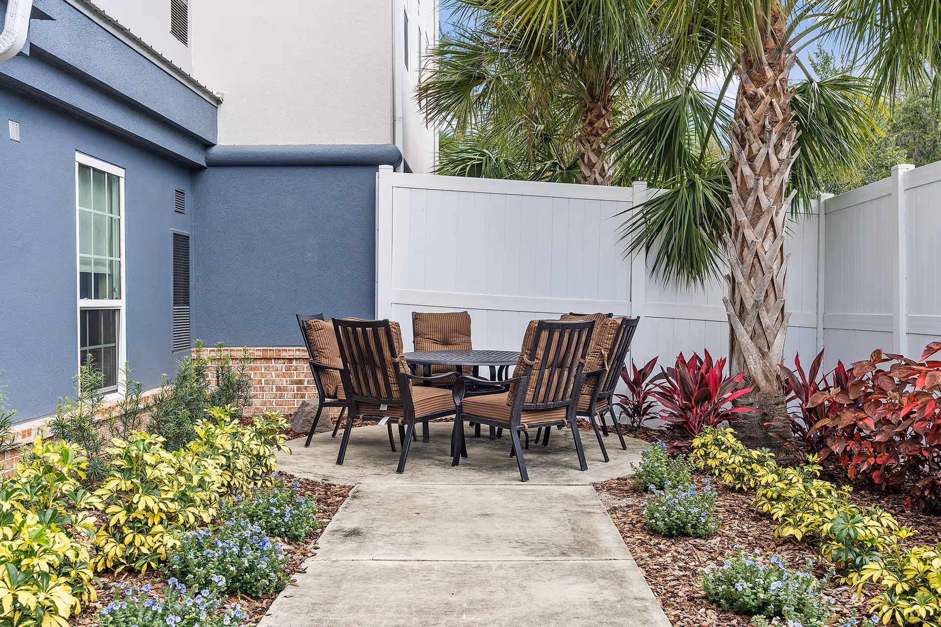 Outdoor patio area with a round table and six cushioned chairs surrounded by landscaped garden beds with various plants and palm trees, enclosed by a white fence and adjacent to a building with blue and white walls.