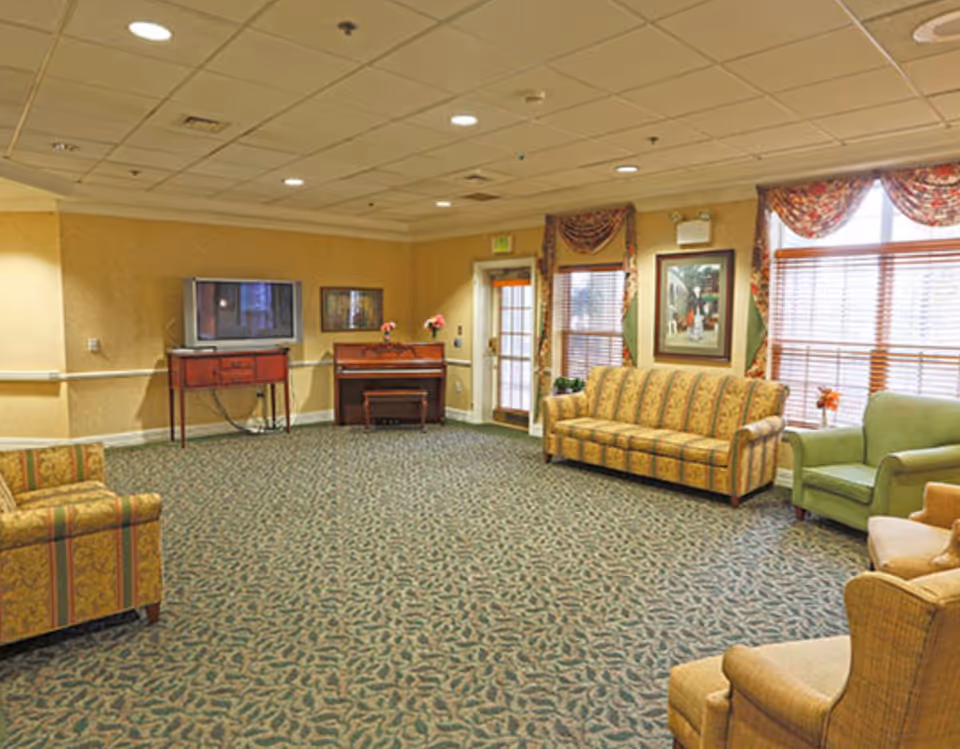 A spacious senior living facility common area with patterned carpet, multiple upholstered chairs and sofas in warm tones, a piano against the wall, a TV on a stand, framed artwork, and windows with floral valances and blinds.