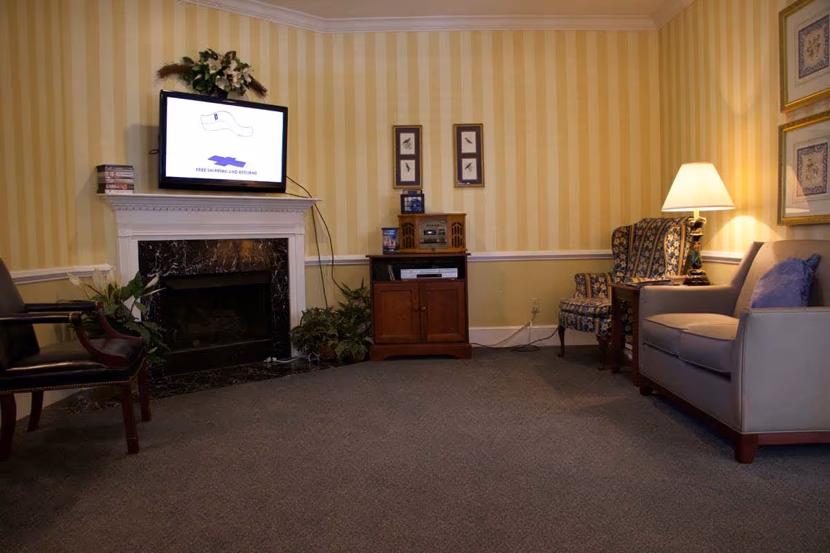 A cozy living room with yellow striped wallpaper, a black marble fireplace with a white mantel, a flat-screen TV mounted above the fireplace, a wooden cabinet with a vintage-style radio, a patterned armchair, a gray sofa with a purple pillow, a side table with a lamp, and framed artwork on the walls.